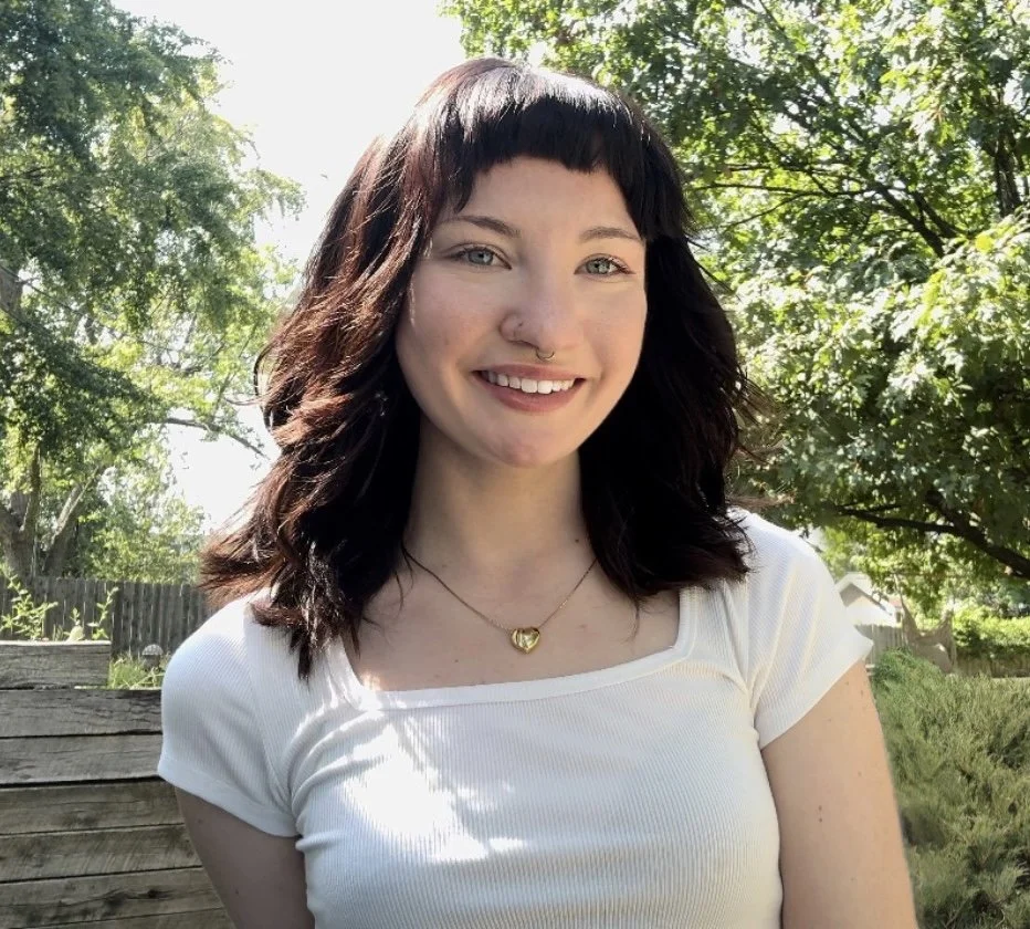 A young woman with shoulder-length dark brown hair and bangs, wearing a white t-shirt and a heart-shaped necklace, standing outdoors in a green, leafy area with sunlight filtering through trees, smiling at the camera.