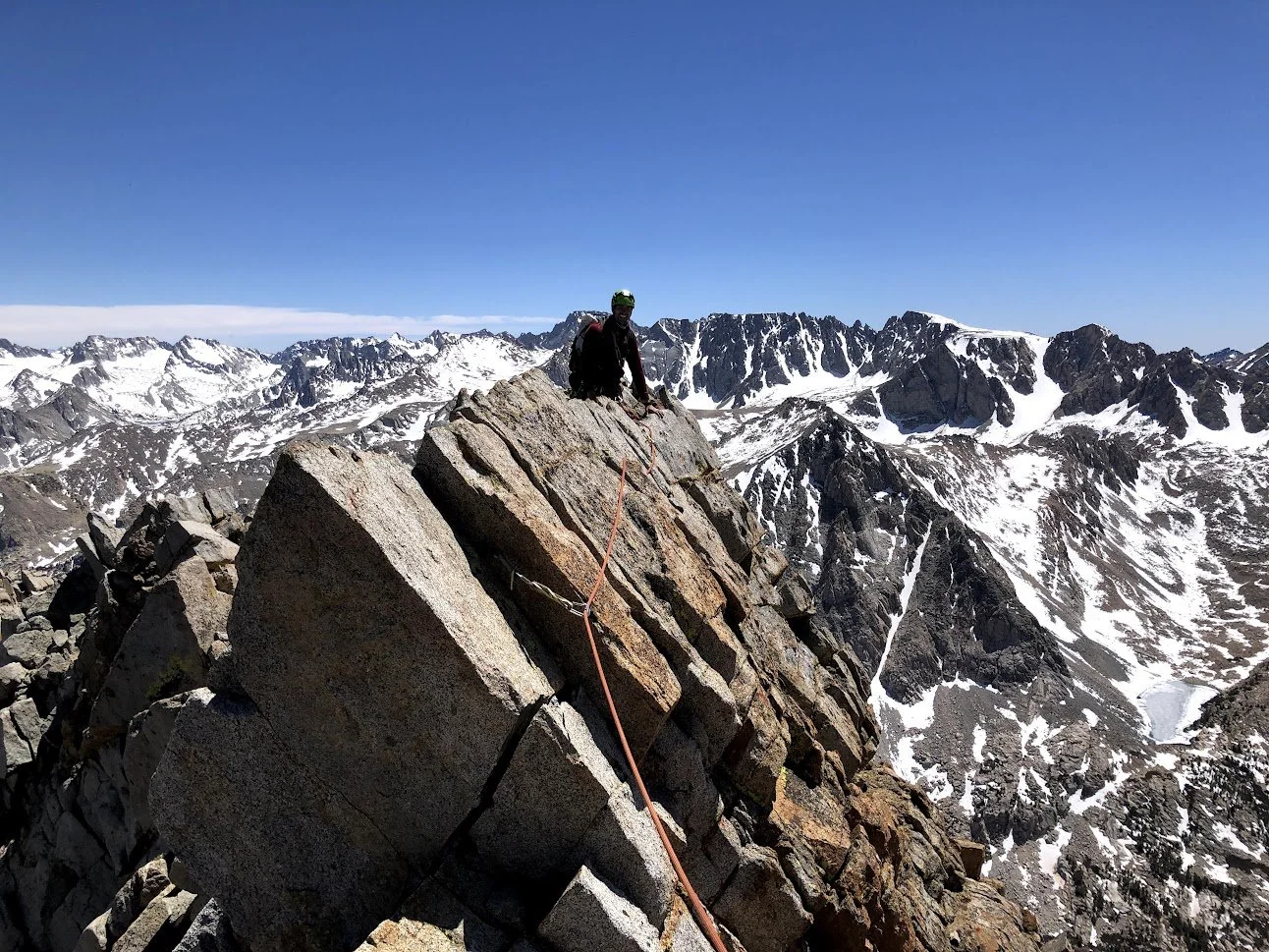 A mountain climber wearing a helmet and backpack scaling a steep rock face with snow-covered peaks in the background under a clear blue sky.