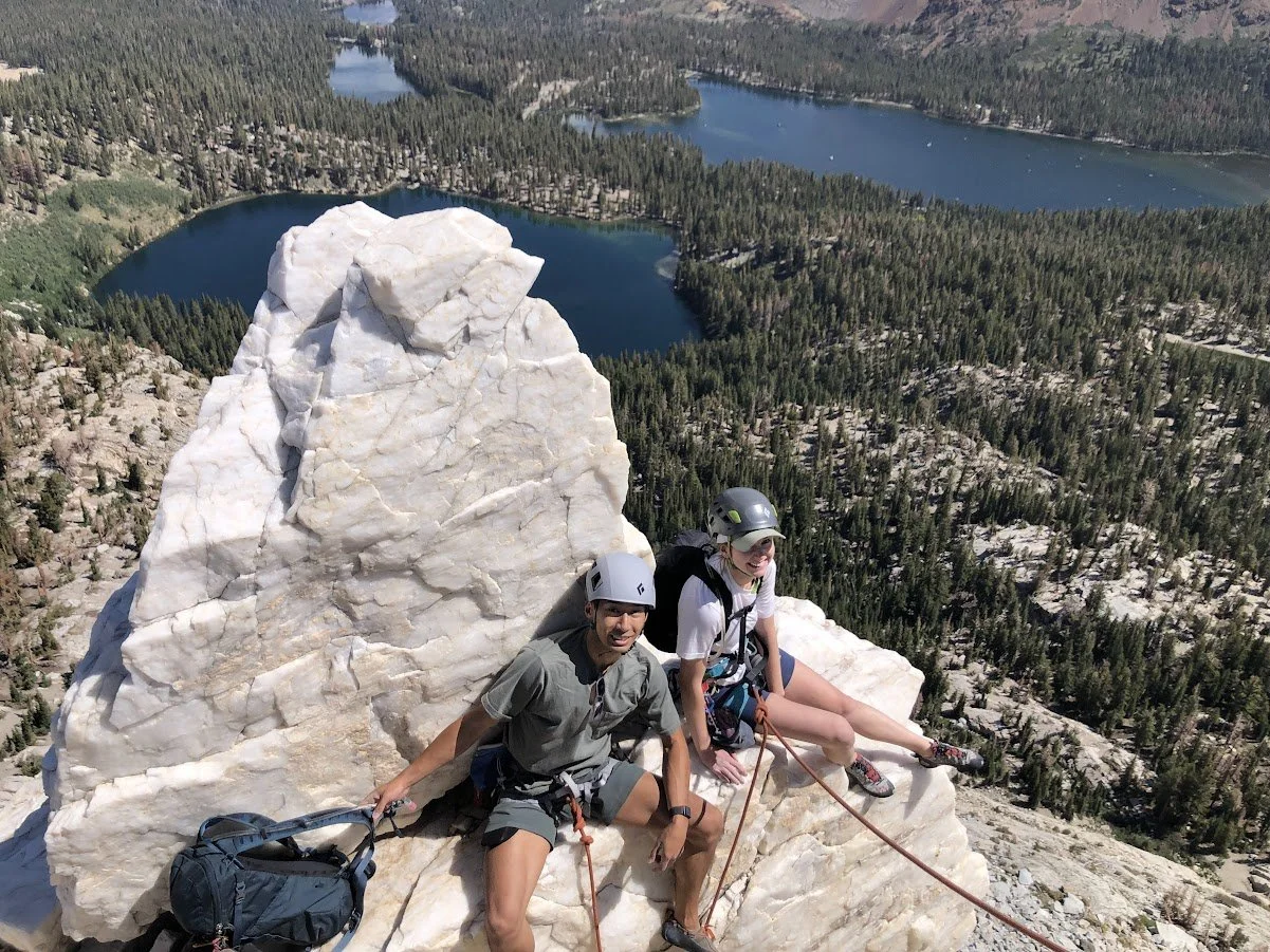 Two climbers sitting on a large white rock on a mountain, with a mountain lake and forest in the background.