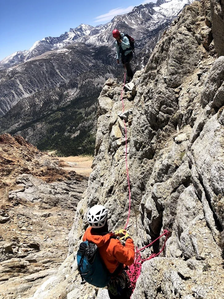 Two climbers, one above and one below, on a steep rocky mountain face, secured with pink climbing ropes and helmets, with snow-capped mountains in the background.