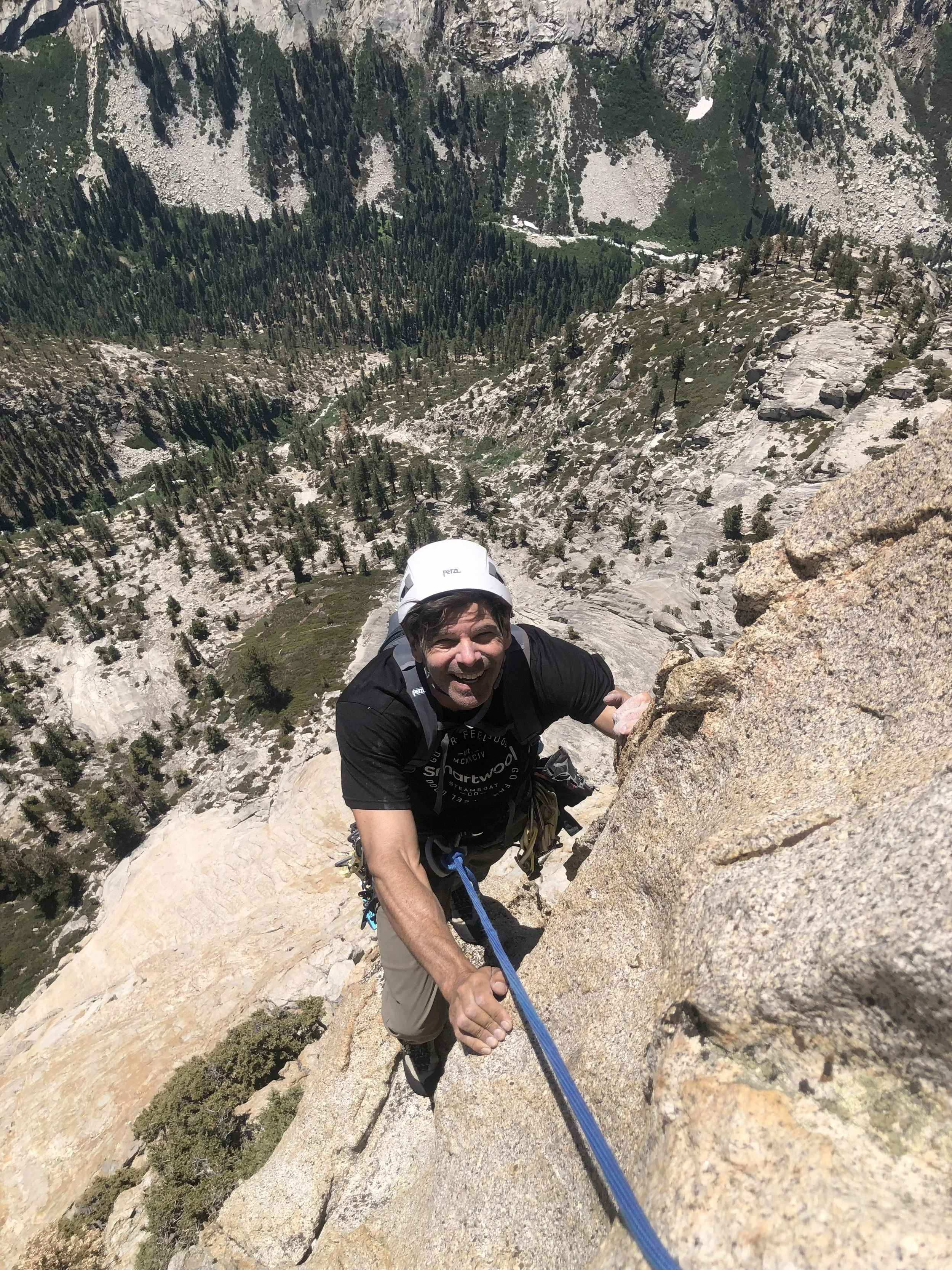 A man wearing a helmet, climbing gear, and a black shirt is ascending a steep rocky mountain face while holding onto a safety rope, with a scenic landscape of trees and mountains in the background.