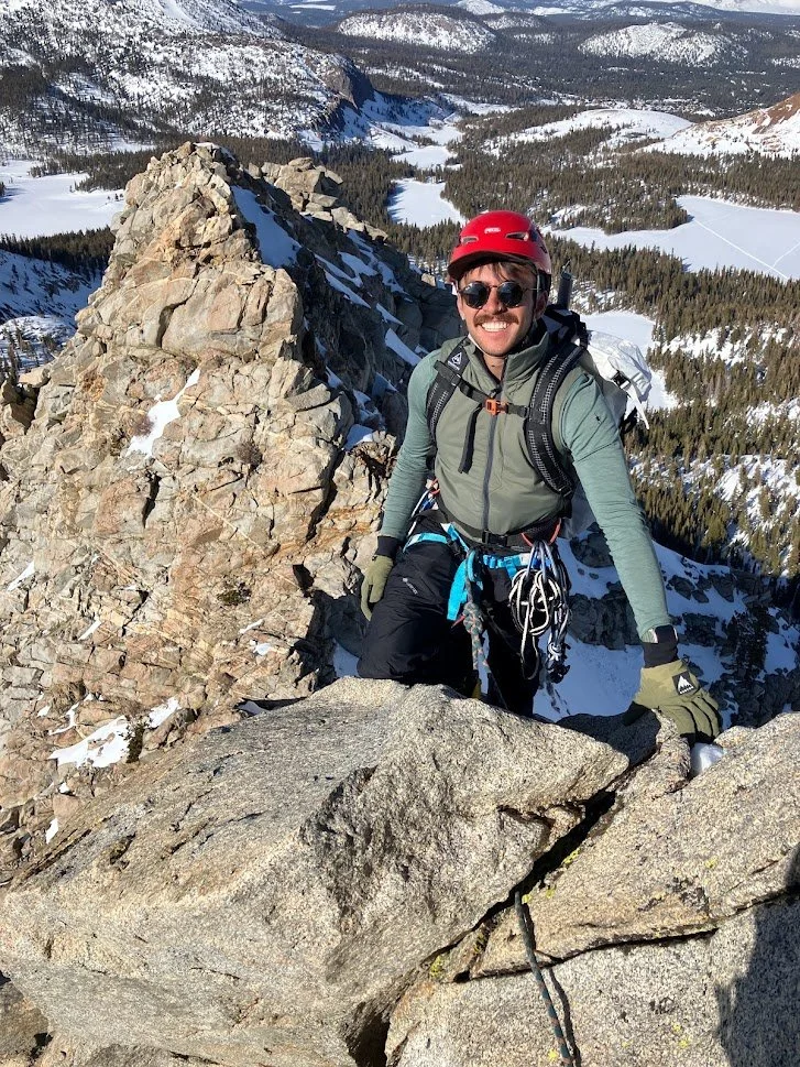 A man in climbing gear, wearing a red helmet and sunglasses, is smiling while climbing rocky mountain terrain in a snowy landscape with trees and mountains in the background.