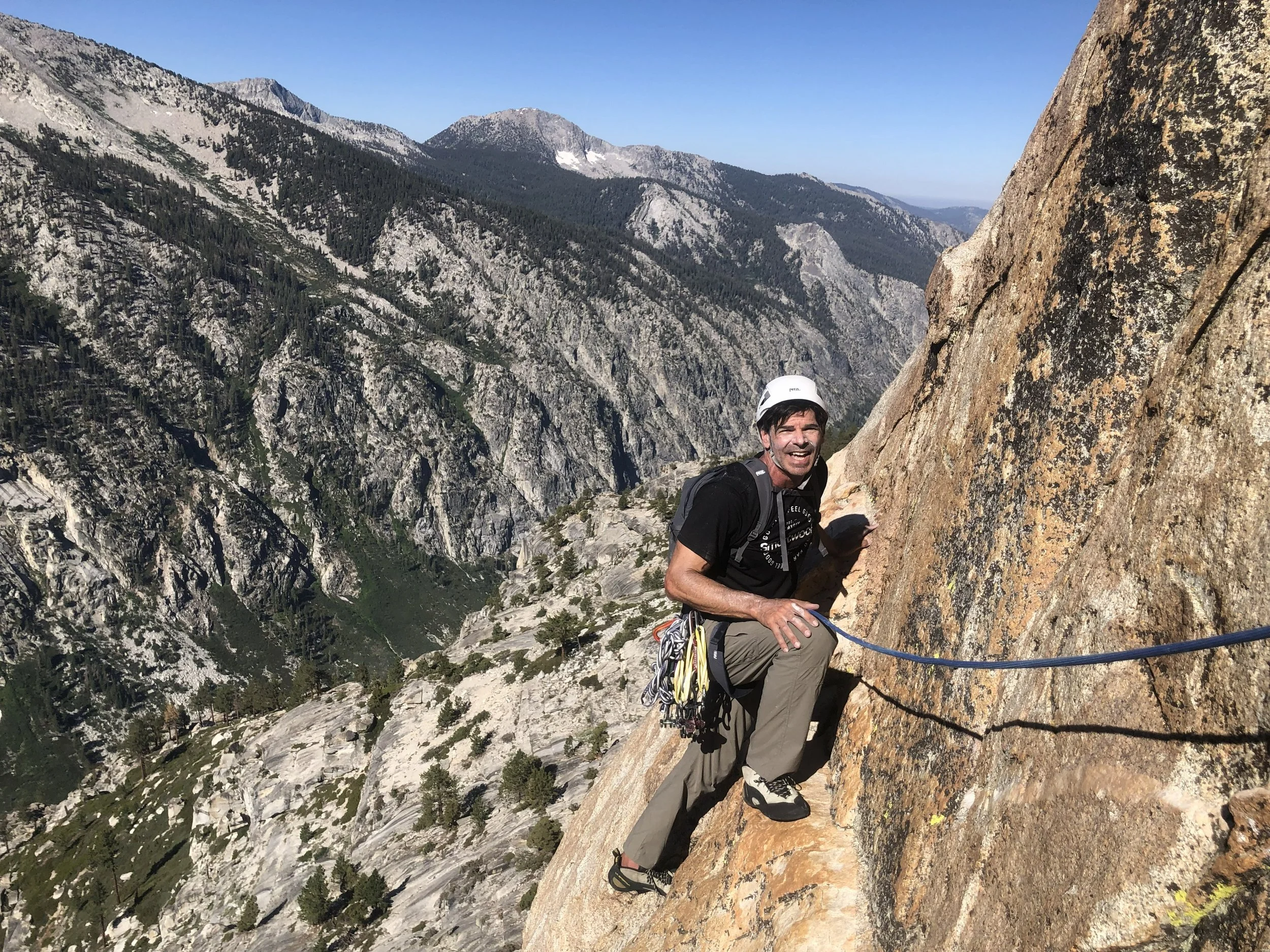 A man with dark hair, wearing a helmet and climbing gear, is rock climbing on a steep mountain face with a rugged mountain landscape in the background.