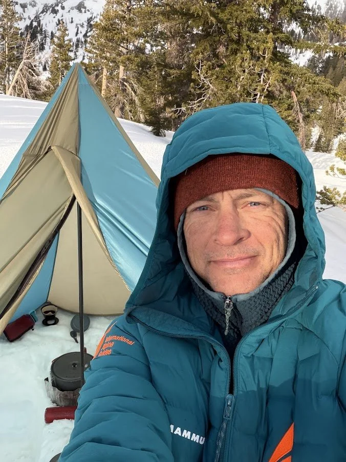 Man taking a selfie outdoors in snowy forest, dressed in blue winter gear with hood, next to a blue and beige tent.