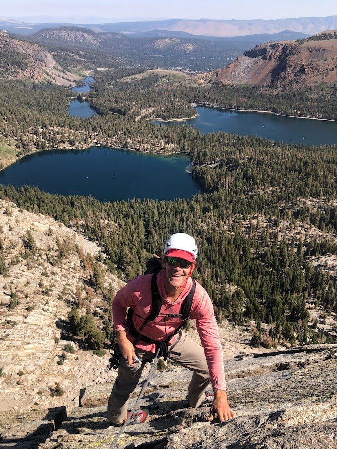 A man climbing a mountain with a scenic view of lakes and forested mountains in the background.