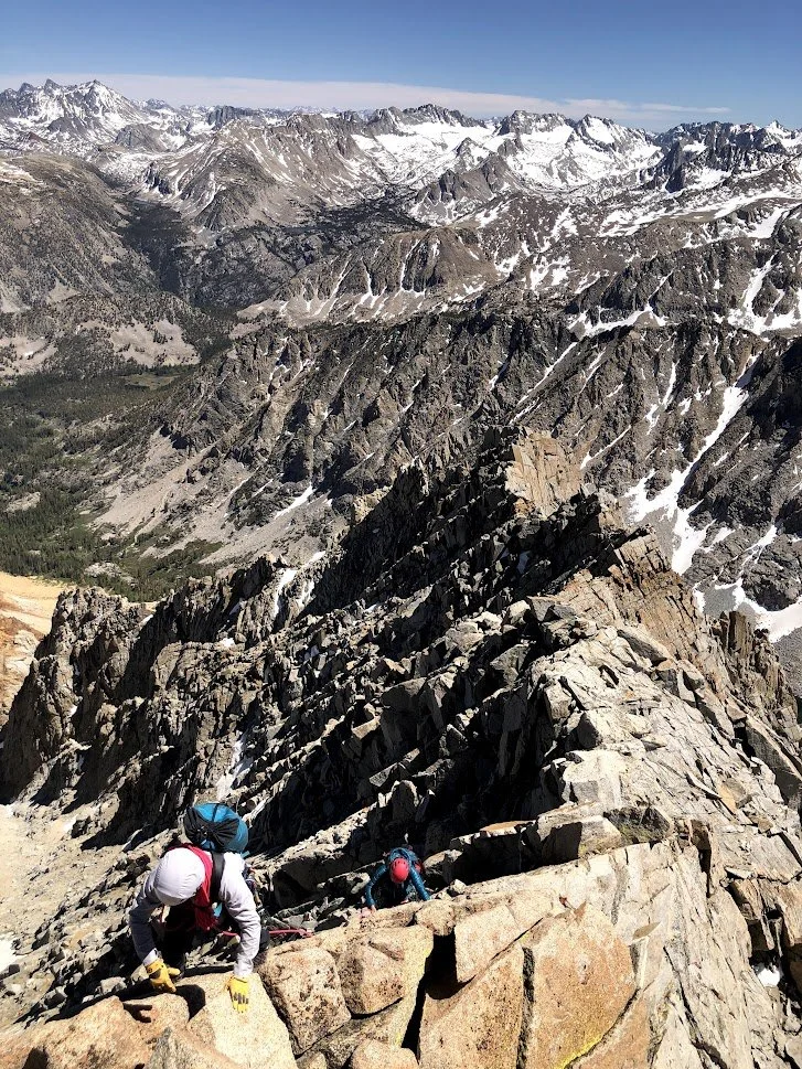 Two climbers in mountain gear ascending a rocky ridge on a mountain range with snow-capped peaks in the distance.