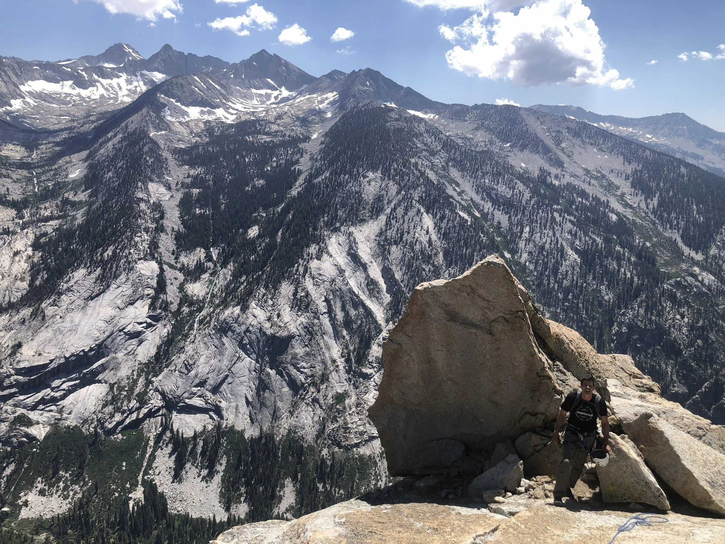 A person hiking on a rocky mountain trail with snow-covered peaks and cliffs in the background, under a partly cloudy sky.