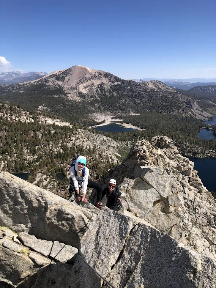 Two climbers with helmets ascending a rocky mountain, with a scenic view of forests, lakes, and mountains in the background under a clear blue sky.