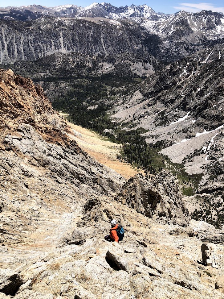 A person hiking up a rugged mountain trail with snow-capped peaks and a valley in the background.