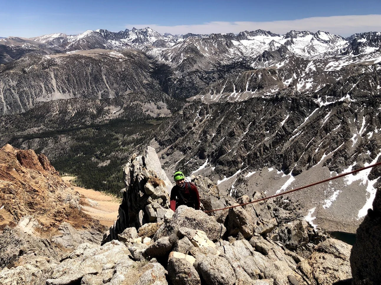 A person wearing a green helmet and red jacket climbing on rocky terrain on a mountain with snow-capped peaks and valleys in the background.