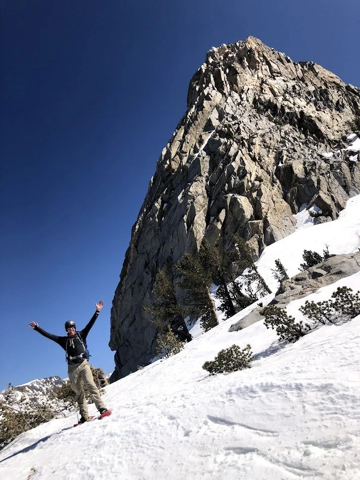 Person standing on snowy mountain slope with arms raised, large rocky mountain and clear blue sky in background.