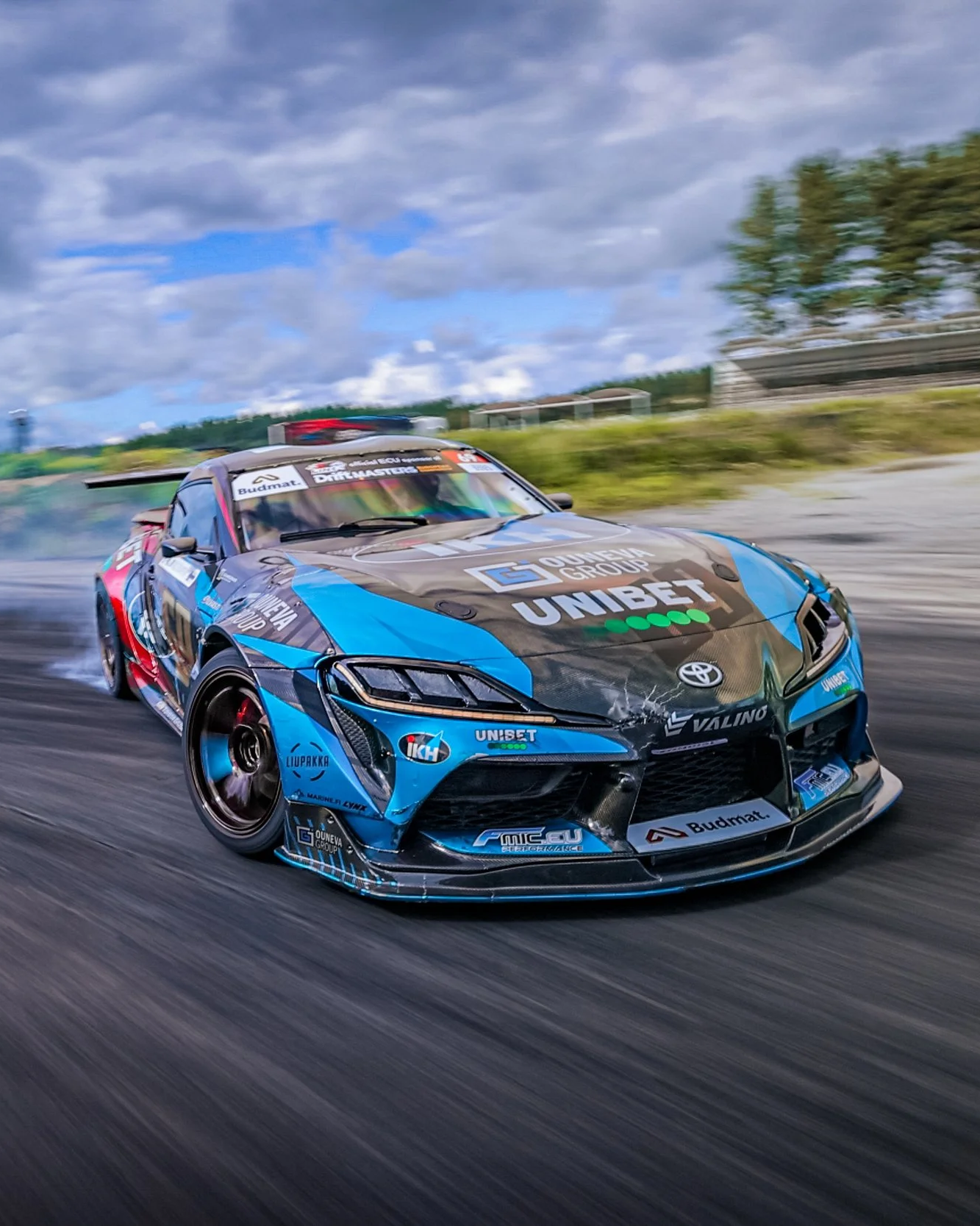 A blue and black race car speeding on a track with clouds in the sky.
