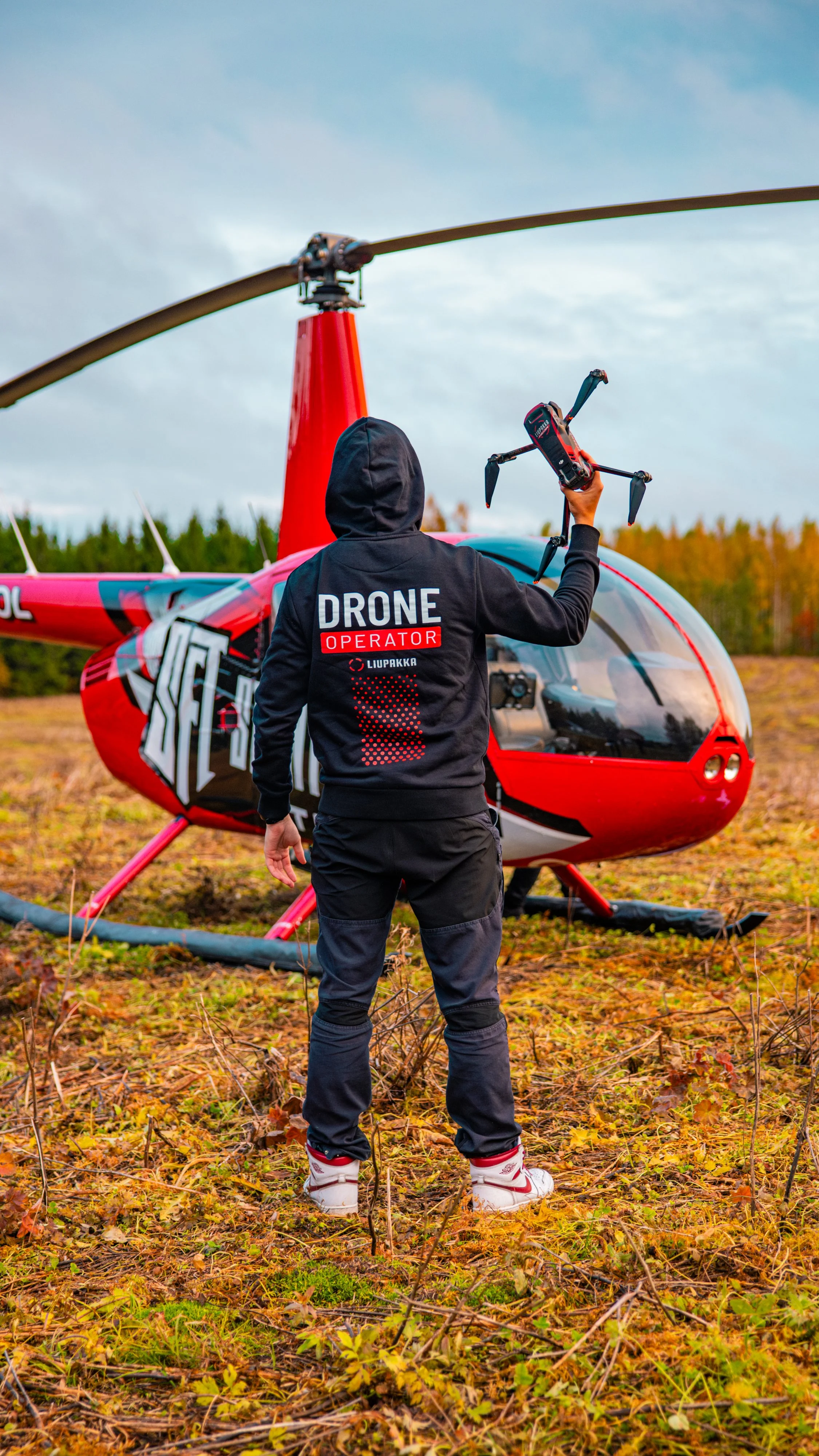Person in a hoodie with 'DRONE OPERATOR' printed on the back holding a drone in front of a red helicopter outdoors on a grassy, autumn landscape.