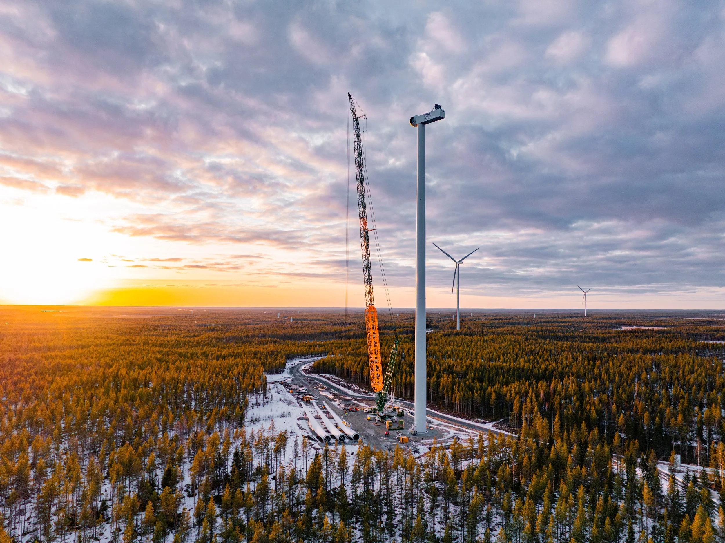 An aerial view of wind turbines and a construction crane in a snow-covered forest at sunset, with a partly cloudy sky.