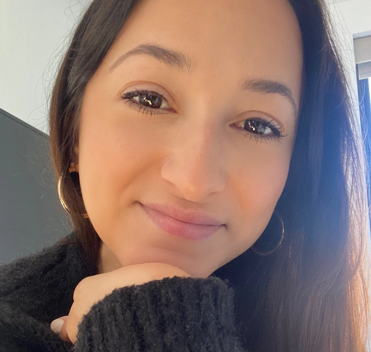 A young woman smiling with a hand resting on her chin, wearing hoop earrings and a black sweater, with natural makeup and long dark hair.