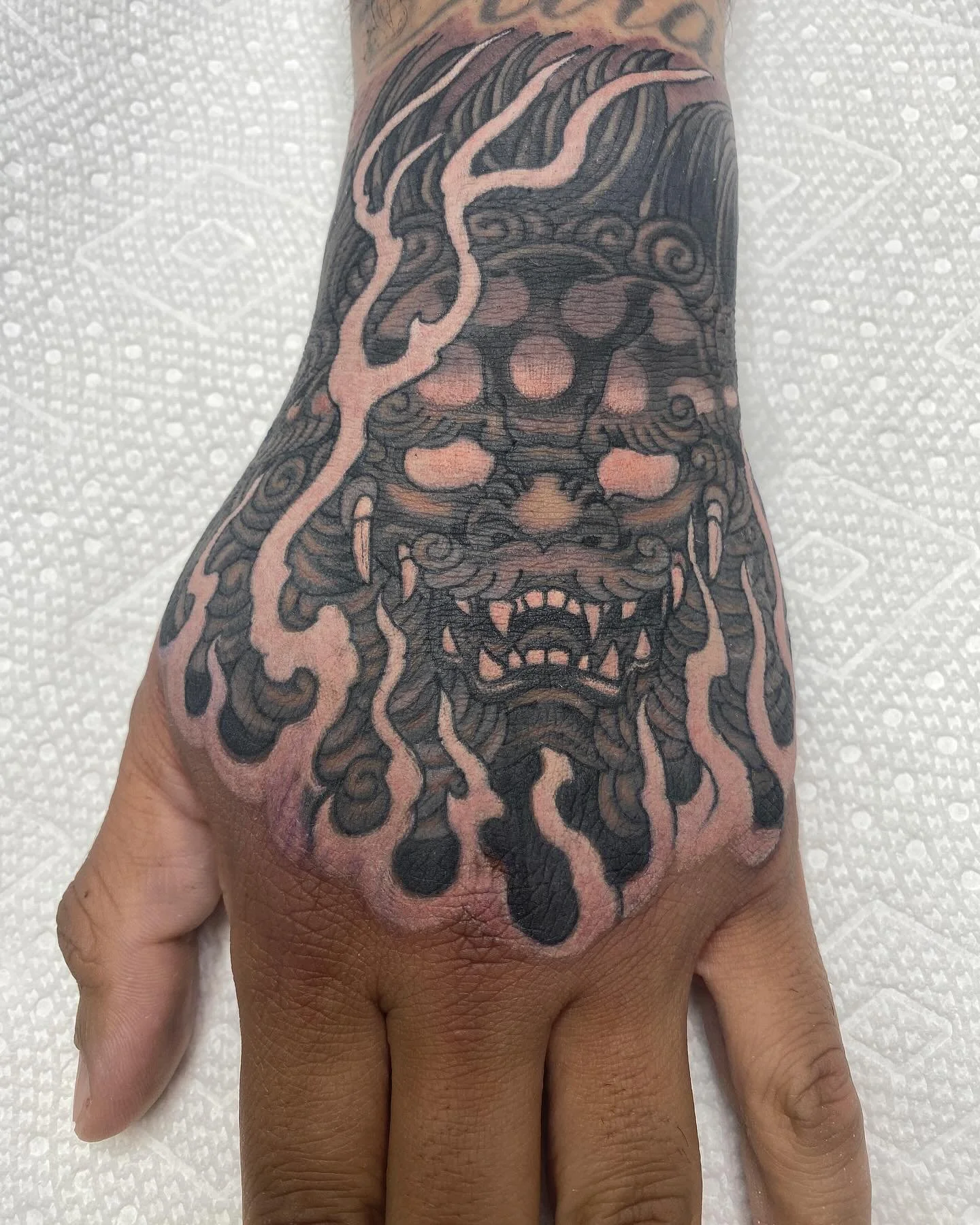 Tattoo of a fierce mythical creature, possibly a demon or oni, with glowing eyes and an open mouth showing sharp teeth, covering the top of a person's hand and extending onto the wrist. The tattoo is in black ink with shading and detailing.
