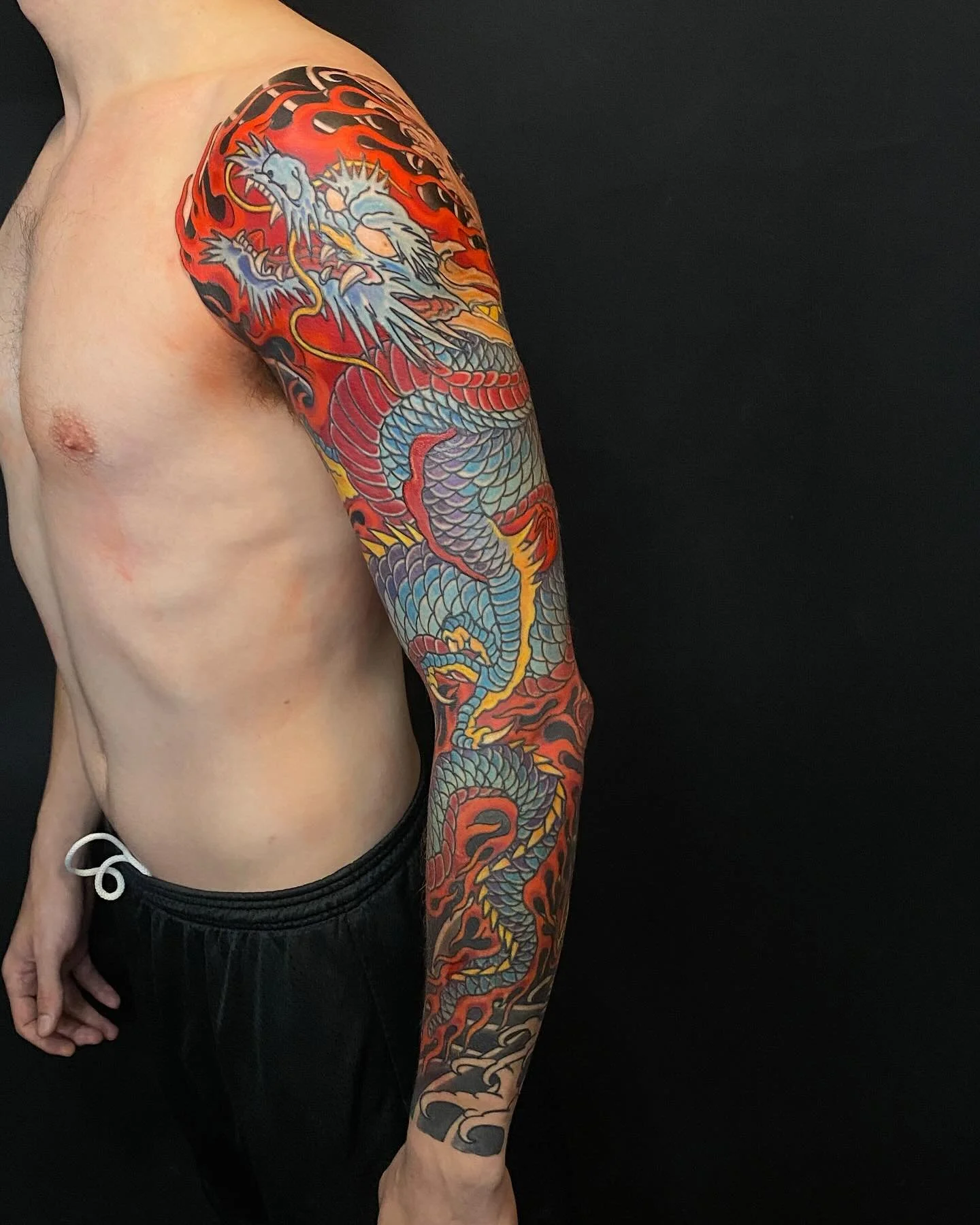 Colorful dragon tattoo covering the person's shoulder and arm, featuring a fierce dragon with detailed scales, surrounded by flames on a black background.