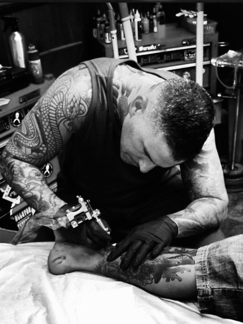 A tattoo artist working on a person's leg in a tattoo shop. The artist is focused, wearing gloves, and using a tattoo machine. The shop has tattoo supplies on shelves in the background.