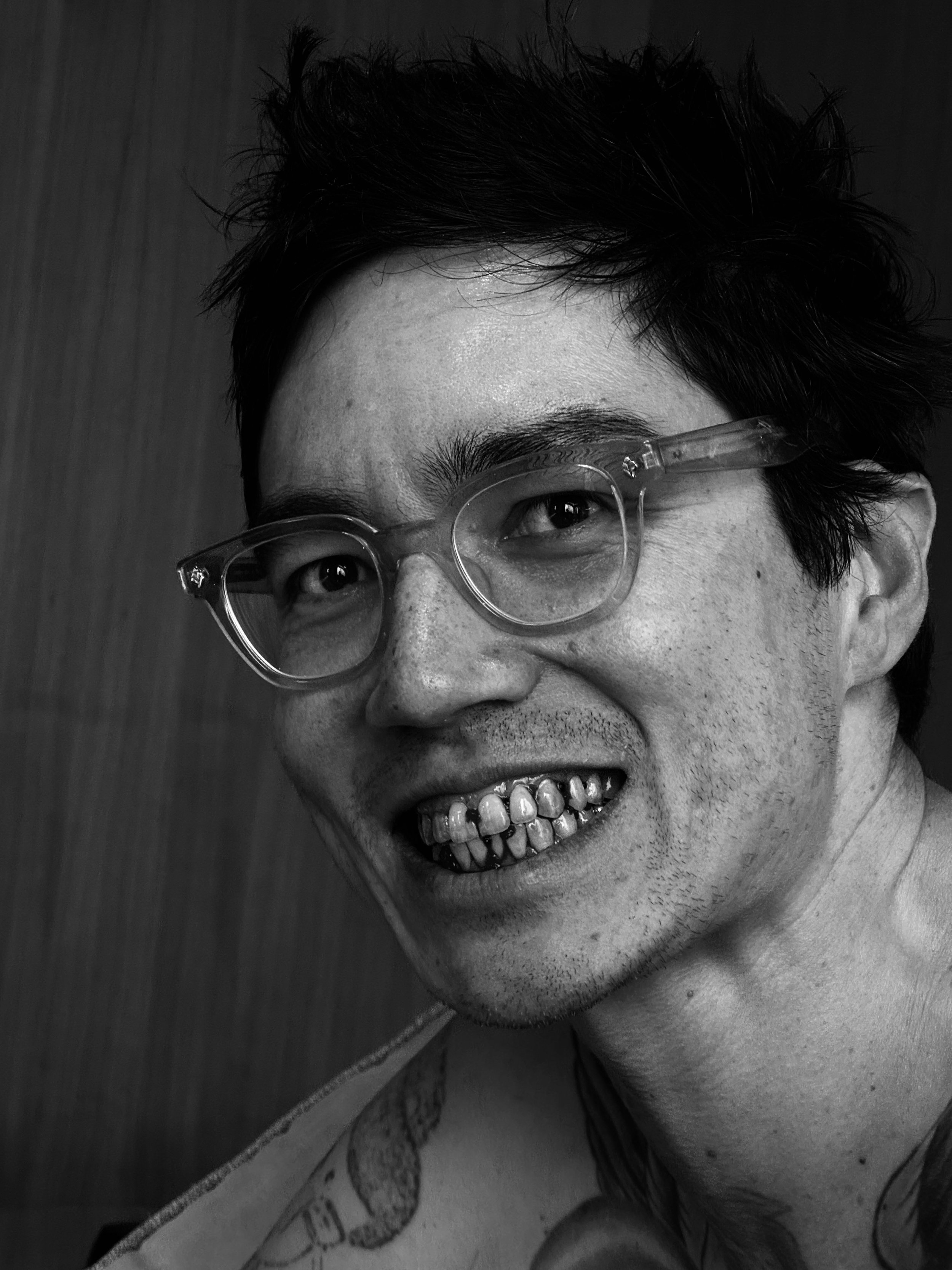 Black and white photo of a smiling Asian man with short dark hair, wearing clear glasses, showing his teeth with some gold and dental work.