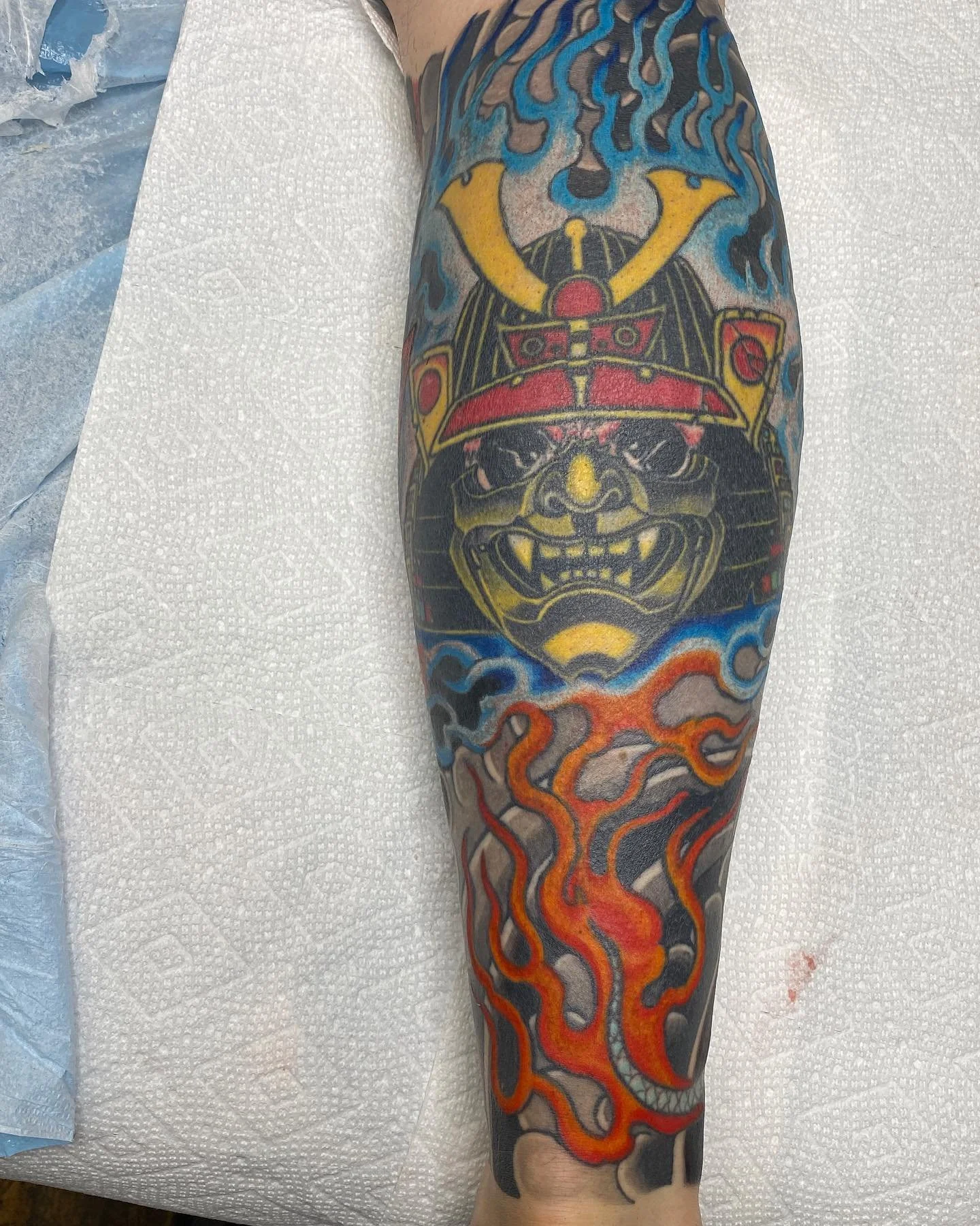 Tattoo of a fierce demon-like face with a horned helmet, surrounded by flames and swirling smoke.