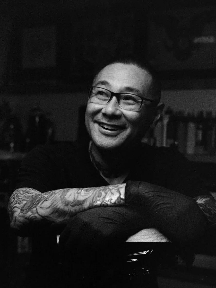 Steven Huie with glasses smiling and resting his chin on crossed arms, showing tattoos on his left arm, in a dimly lit room.