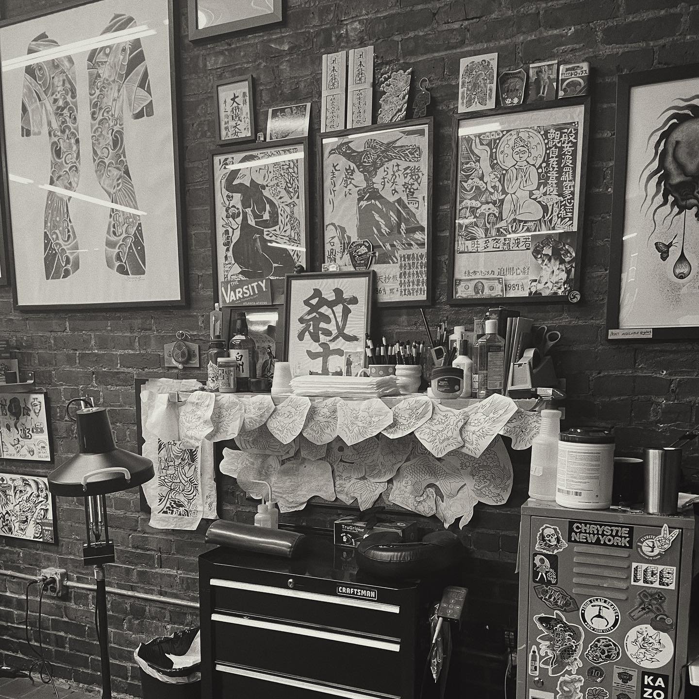 A cluttered workspace against a brick wall with various framed art, posters, and decorative items, a small table covered with a cloth holding bottles and art supplies, a black Craftsman toolbox, and a collection of stickers on a nearby object.