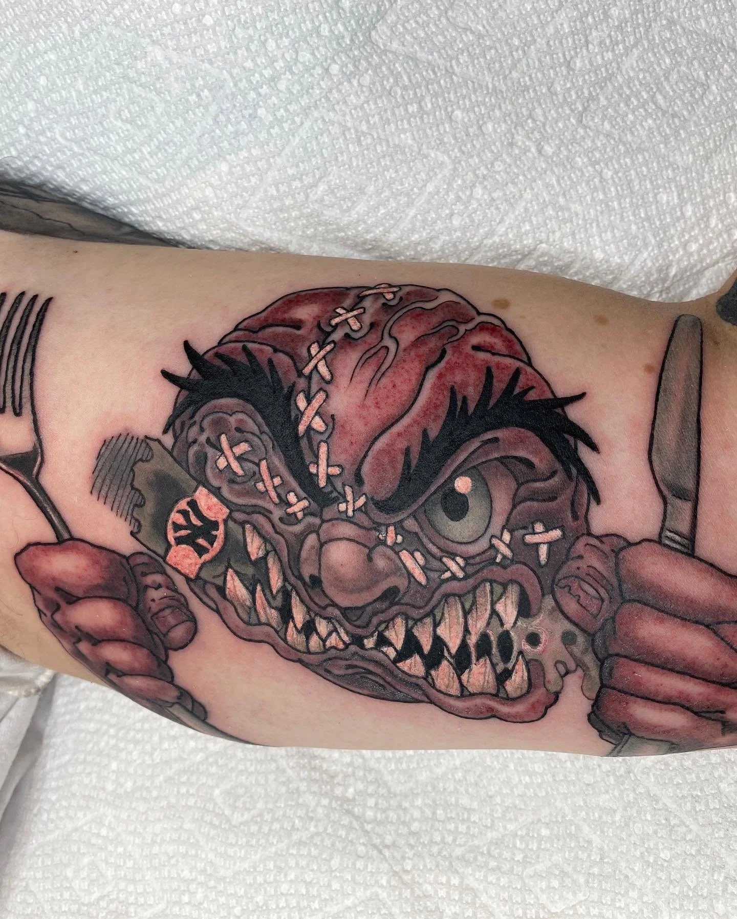 Tattoo of a zombie-like creature with stitches across its head, holding a fork and knife, with red and gray tones, on a person's arm.