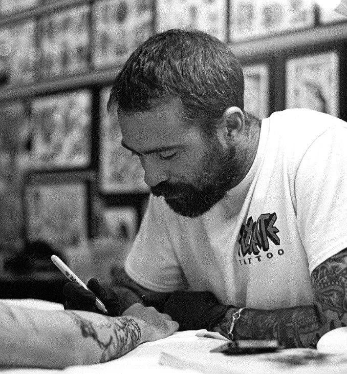 A man with a beard and tattoos getting a tattoo on his arm in a tattoo parlor.