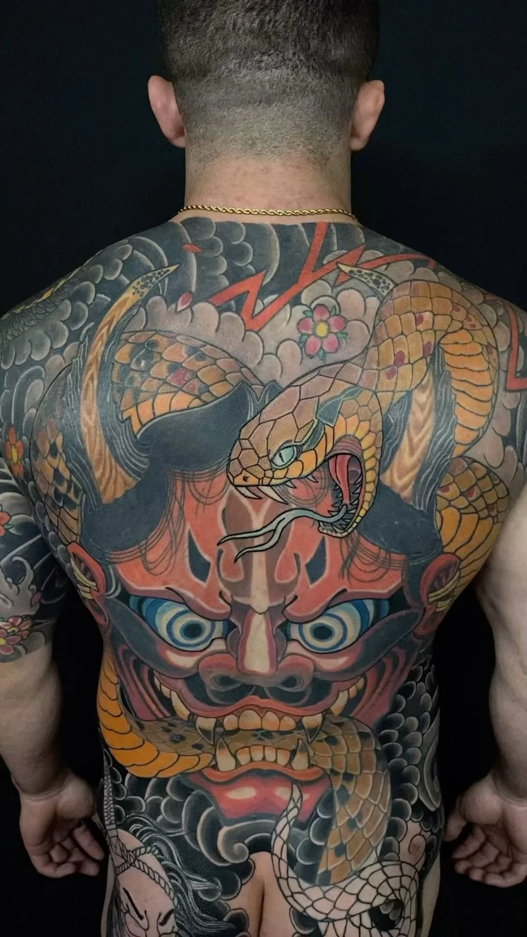 Full back tattoo of a traditional Japanese style mask, a snake, clouds, roses, and a mountain, with vibrant colors and detailed linework.