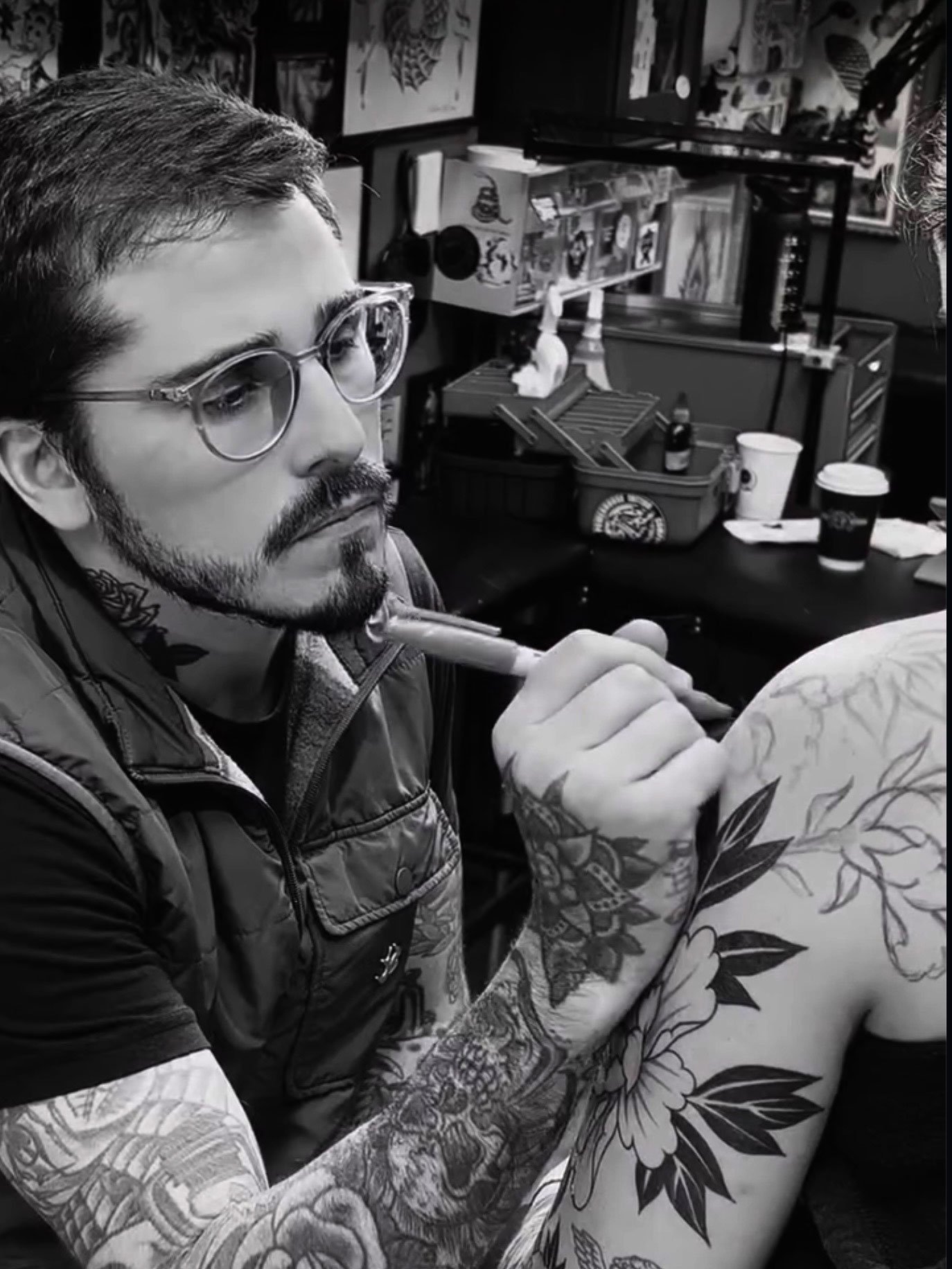 A man with glasses and tattoos drawing on another person's shoulder in an art studio.