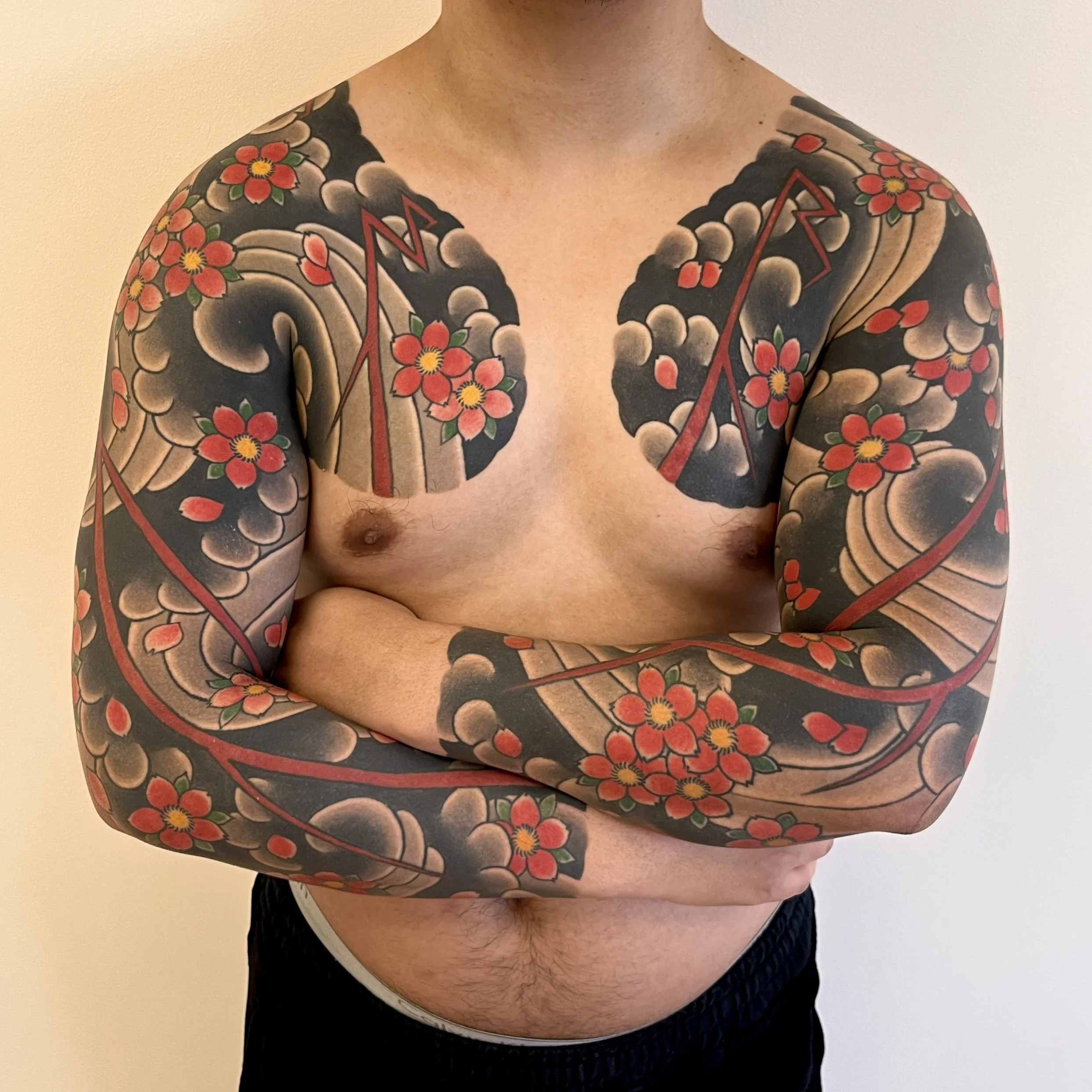 A person with extensive traditional Japanese-style tattoos featuring cherry blossoms, cloud patterns, and red lines across both arms and shoulders, standing against a plain light-colored background.