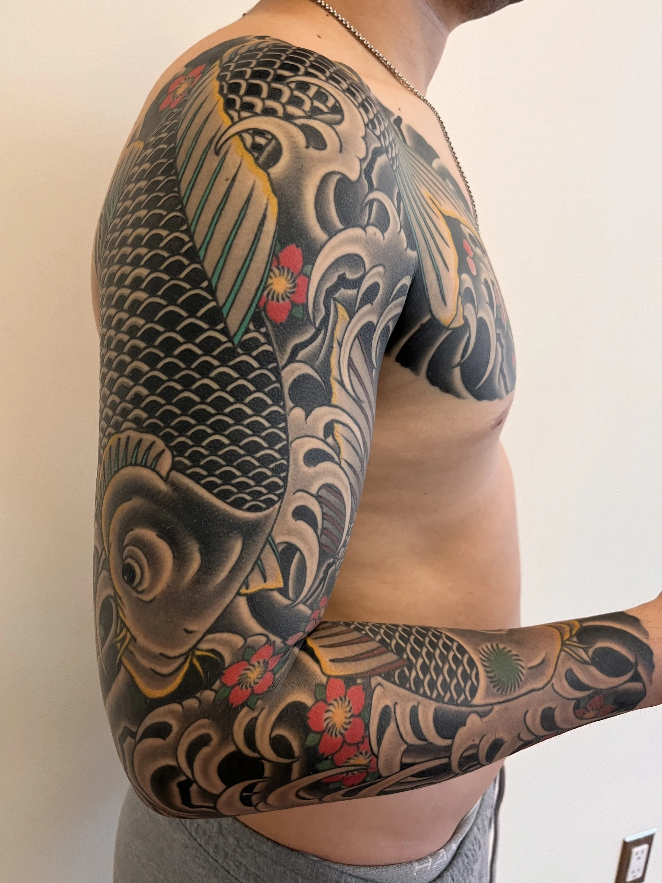 Close-up of a person's arm and shoulder covered in a Japanese-style tattoo featuring waves, koi fish, and red flowers, with a minimal background of a plain white wall.