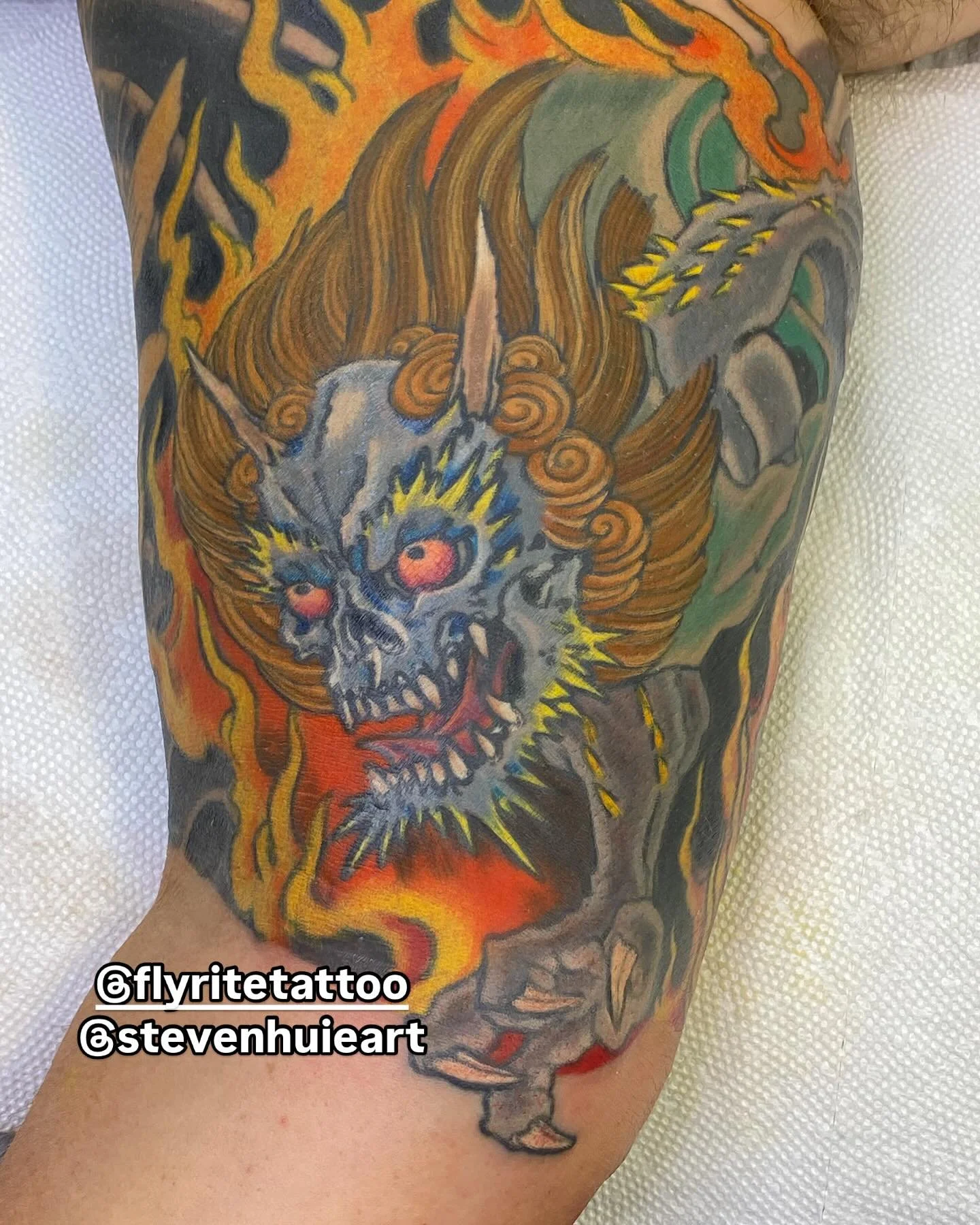 A colorful tattoo of a fantasy creature with a skull face, fiery mane, and horns on a person's arm.