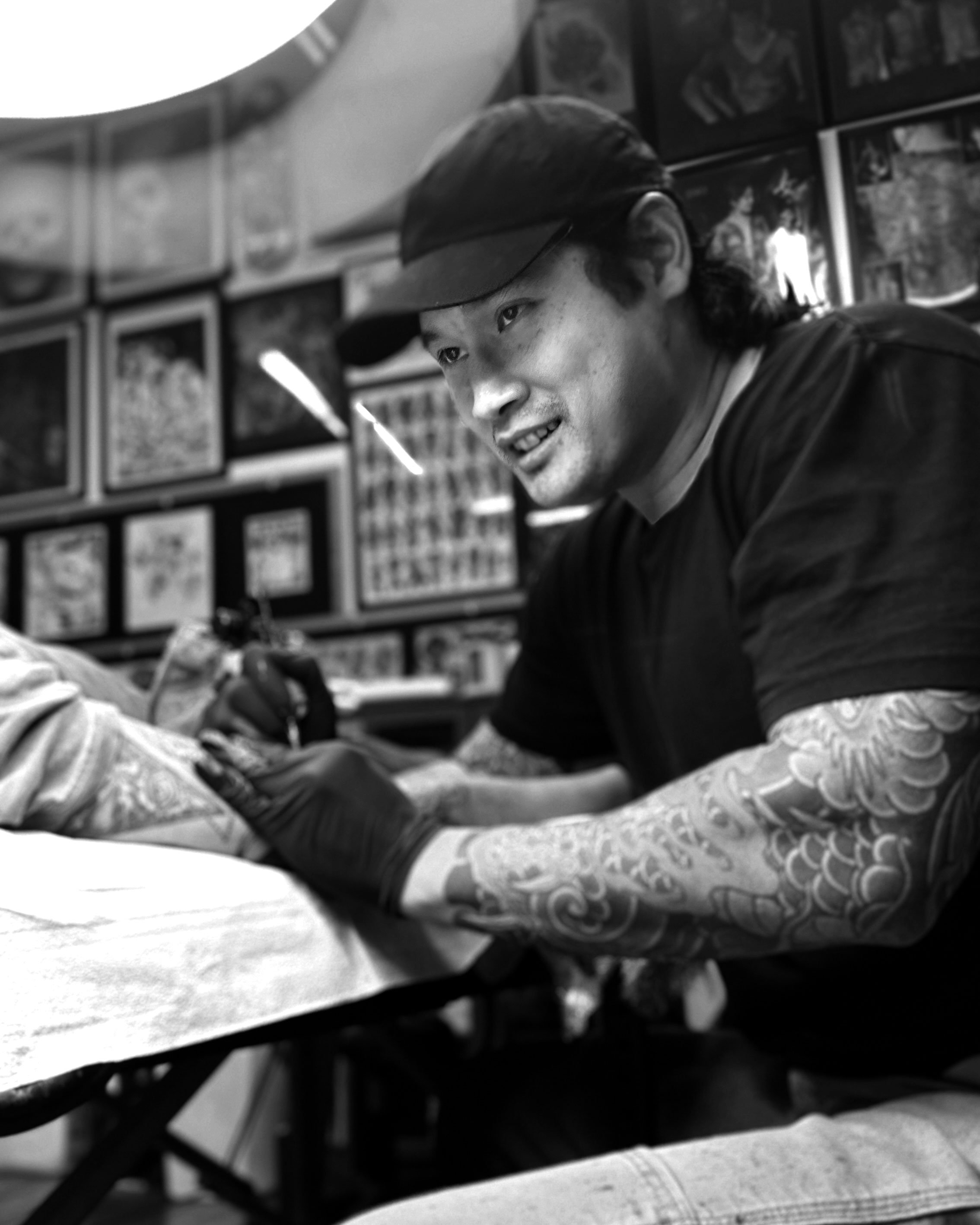 Black and white photo of a woman sitting on a chair, wearing sunglasses and a dark outfit, with a confident posture in an indoor setting.