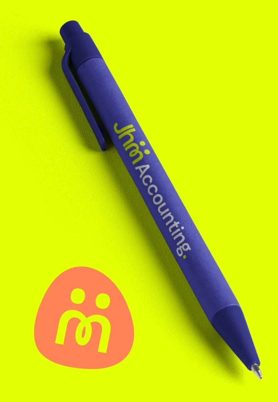 Blue pen on yellow background with the text "Jham: Accountings" printed on it. A pink logo with the letter "M" and two dots next to it is also visible near the pen.