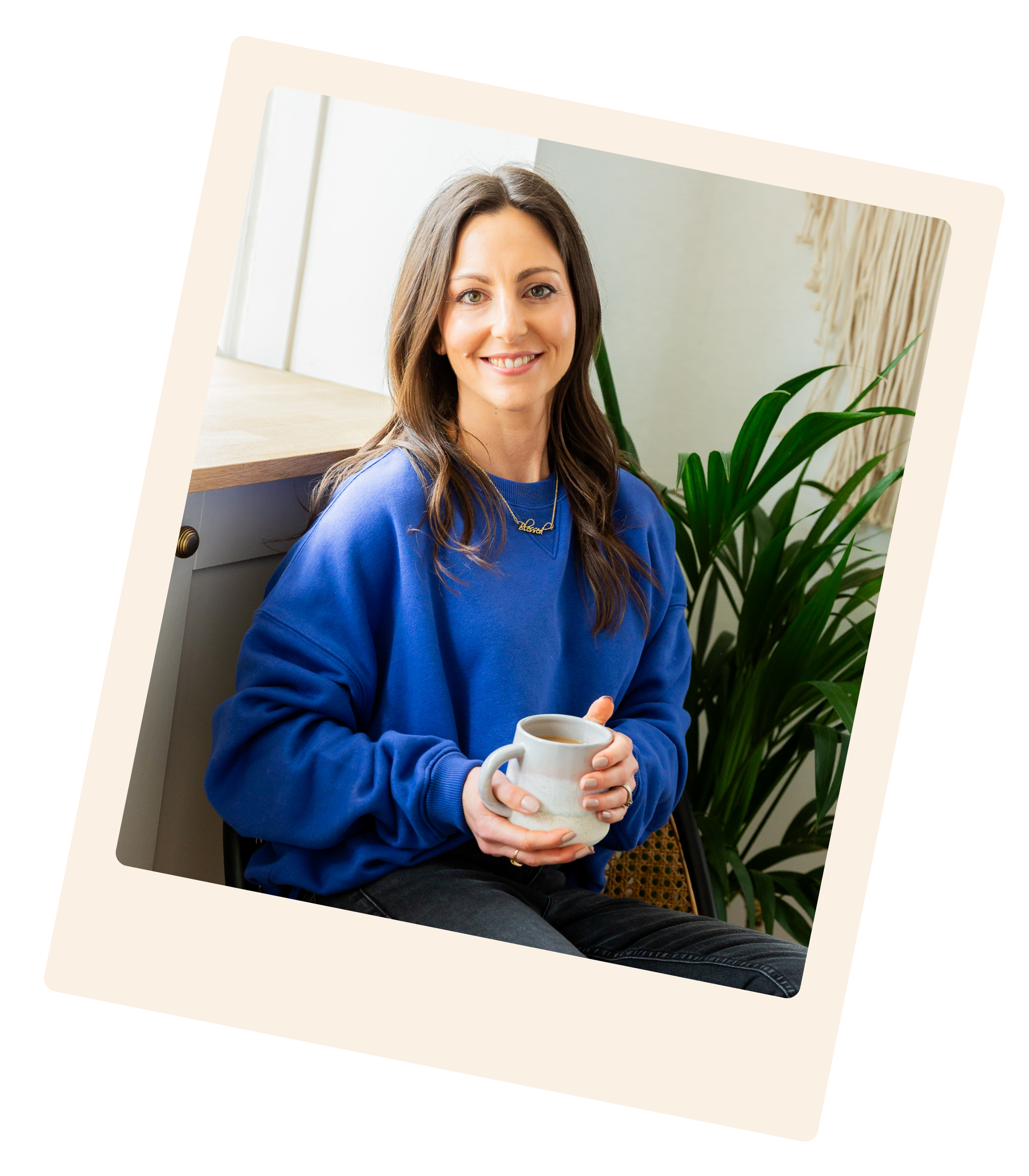 Woman with brown hair smiling, wearing a blue sweatshirt and holding a white coffee mug, sitting indoors near a large green plant, Kent-based brand designer at Lauren Design Studio.