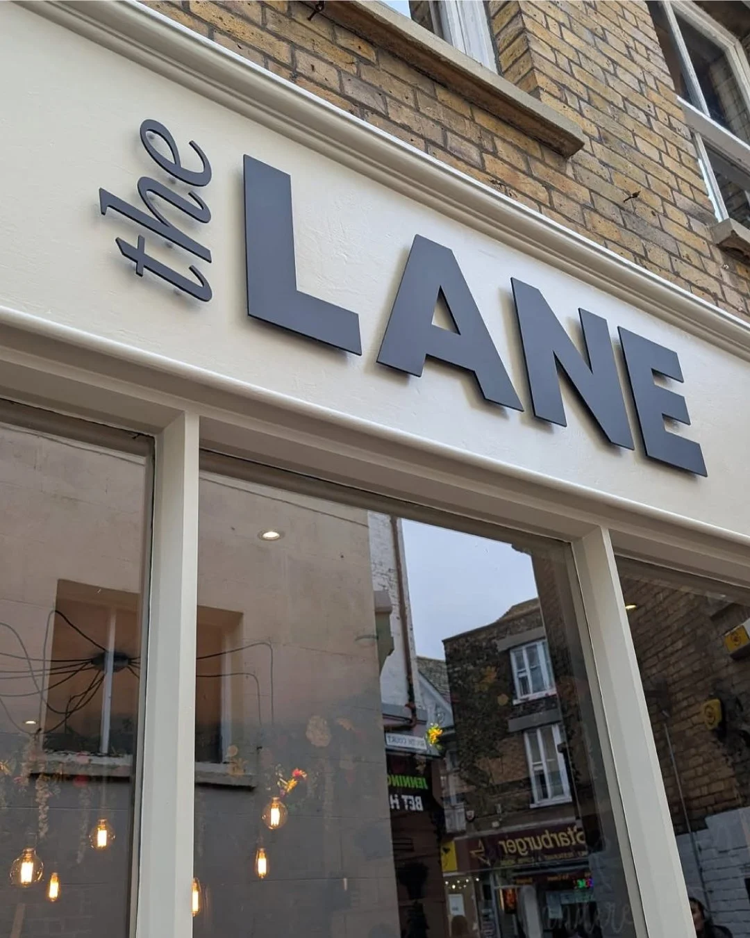 The storefront sign reads 'the' in small letters and 'LANE' in large capital letters above the glass window. The background features a brick building and reflections of nearby shops and street scene.