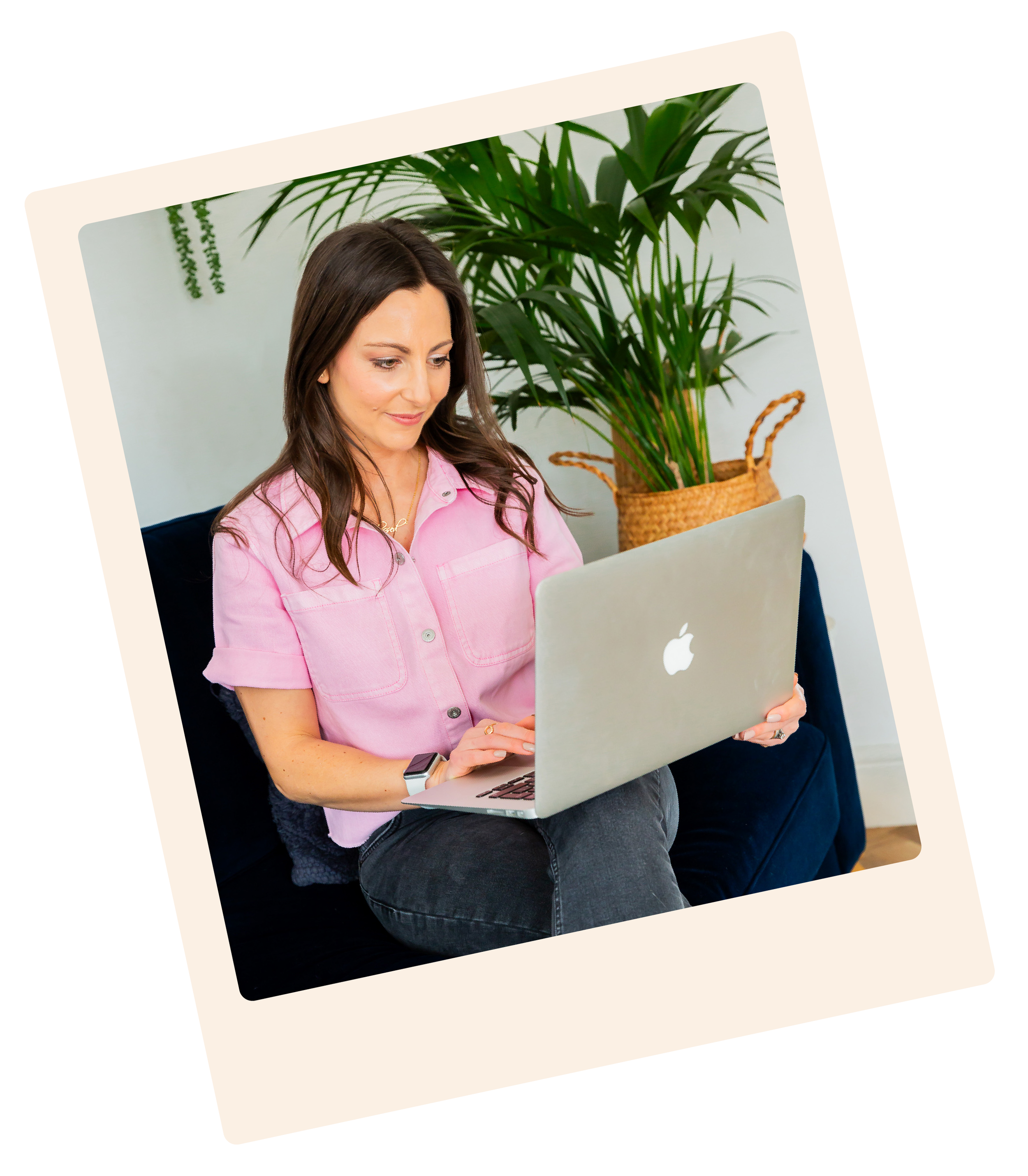 Woman with dark hair wearing pink shirt working on a silver MacBook laptop, sitting on a dark blue couch with a large green potted plant behind her, Kent-based brand designer at Lauren Design Studio.