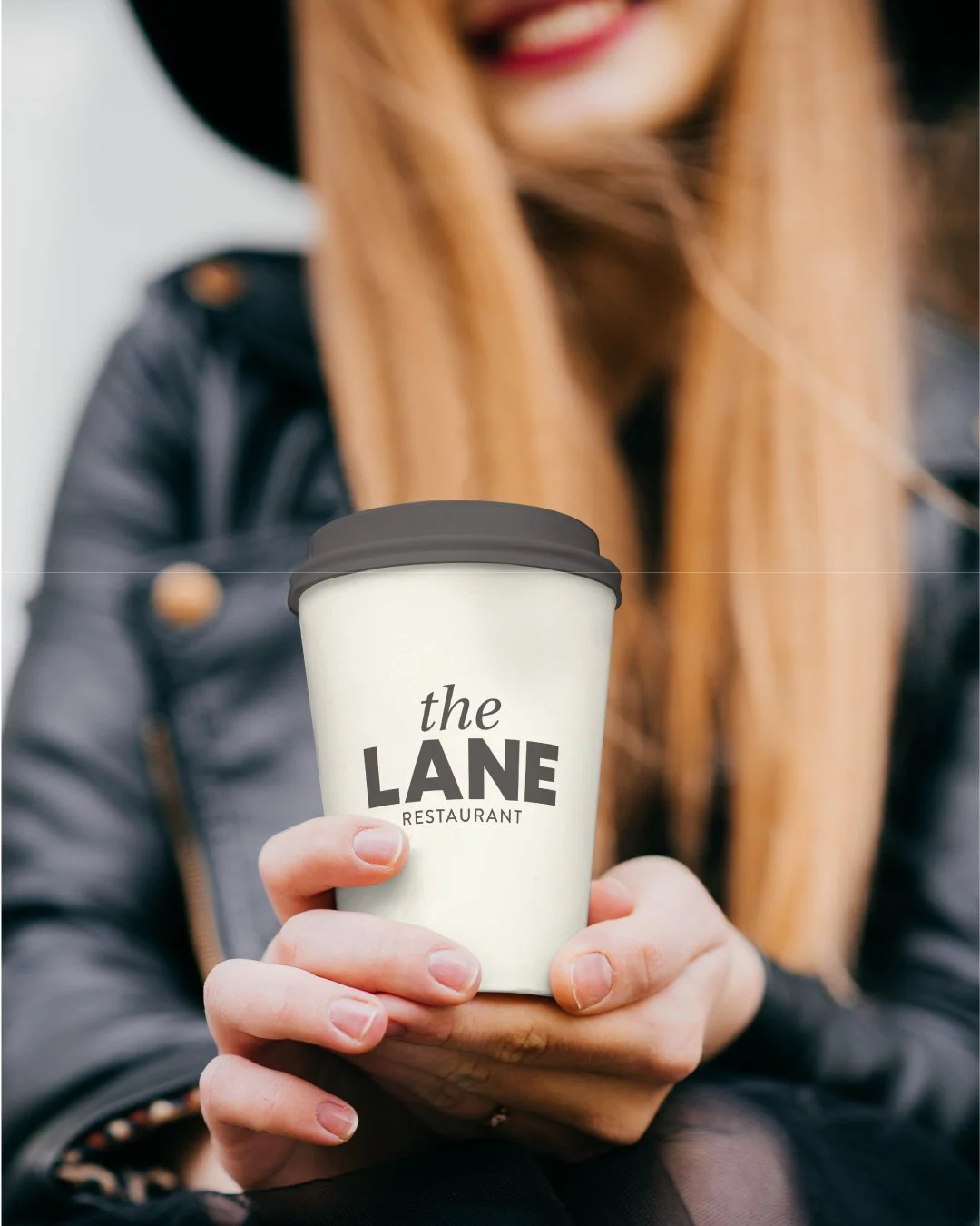 Person holding a coffee cup with 'the LANE RESTAURANT' logo, wearing a black leather jacket and hat, smiling.