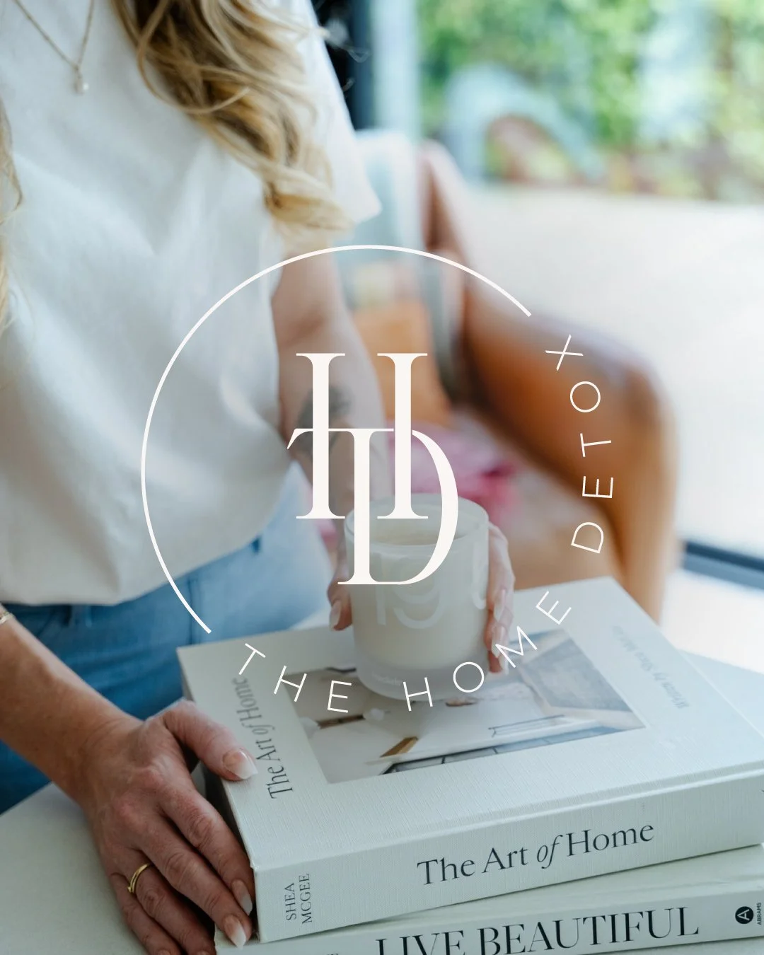 Person holding a white mug over a coffee table with a large book titled 'The Art of Home' and another book titled 'Live Beautiful' on it, with a background of a cozy, well-lit room. Part of the new brand design for The Home Detox