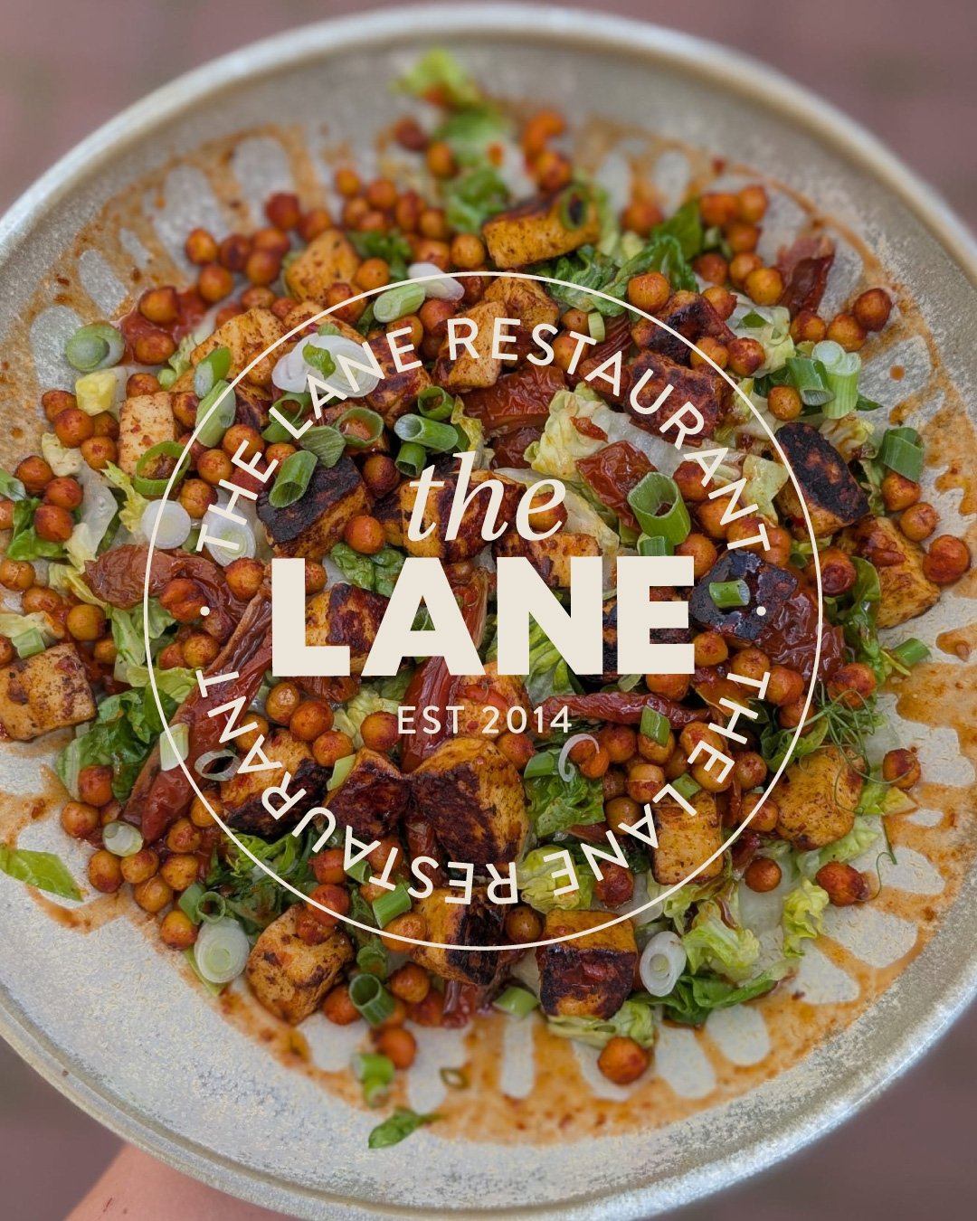 A plate of food with grilled chicken, roasted vegetables, and greens, with a circular overlay of text that reads 'the LANE, the restaurant by the lane, established 2014'.