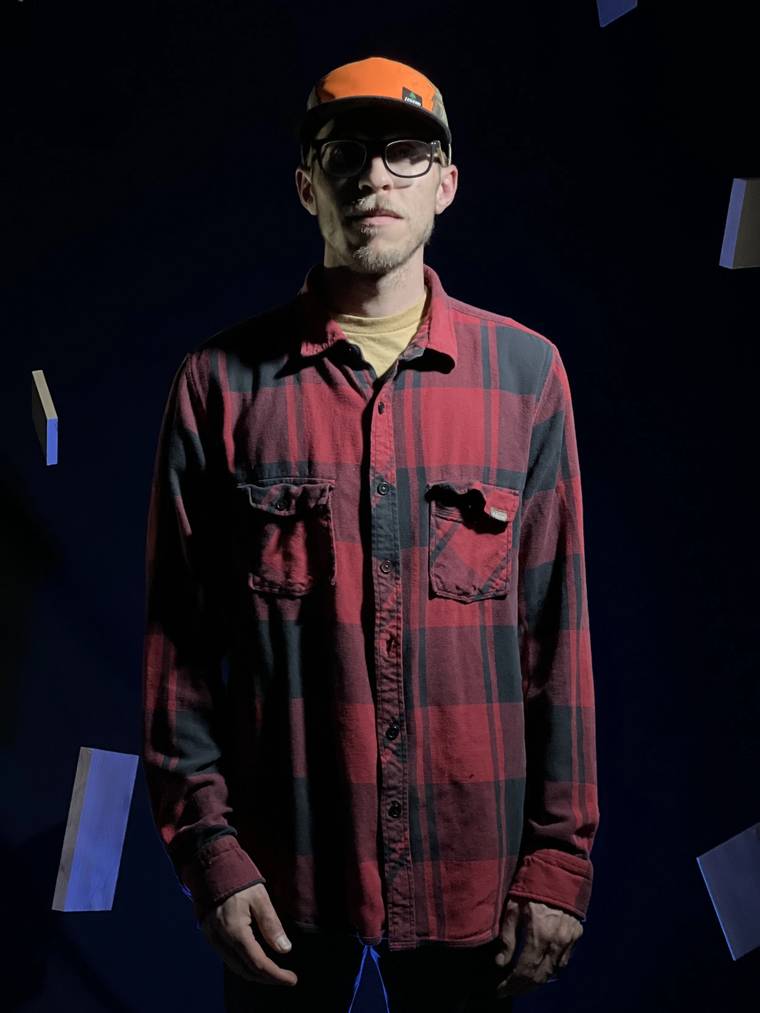 A man wearing glasses, a red and black plaid shirt, and a colorful cap standing against a dark background with floating rectangular objects.