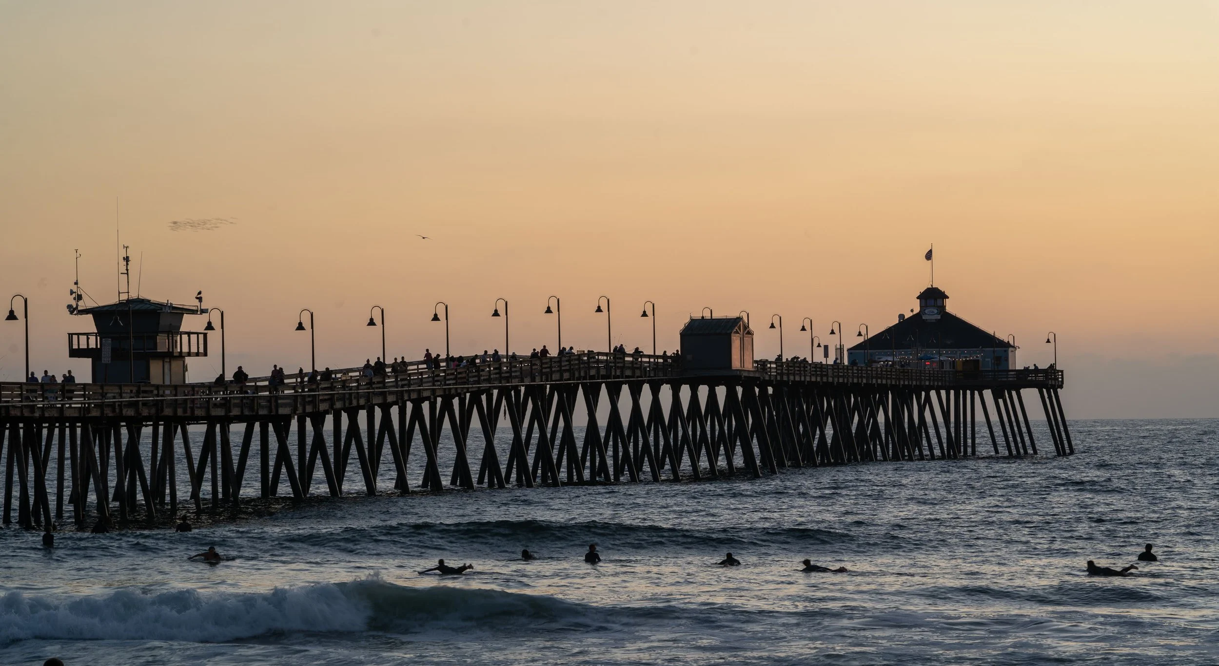 Beach at sunset with a pier extending into the water, people walking on the pier, surfers in the ocean, and a lighthouse on the pier at the end.