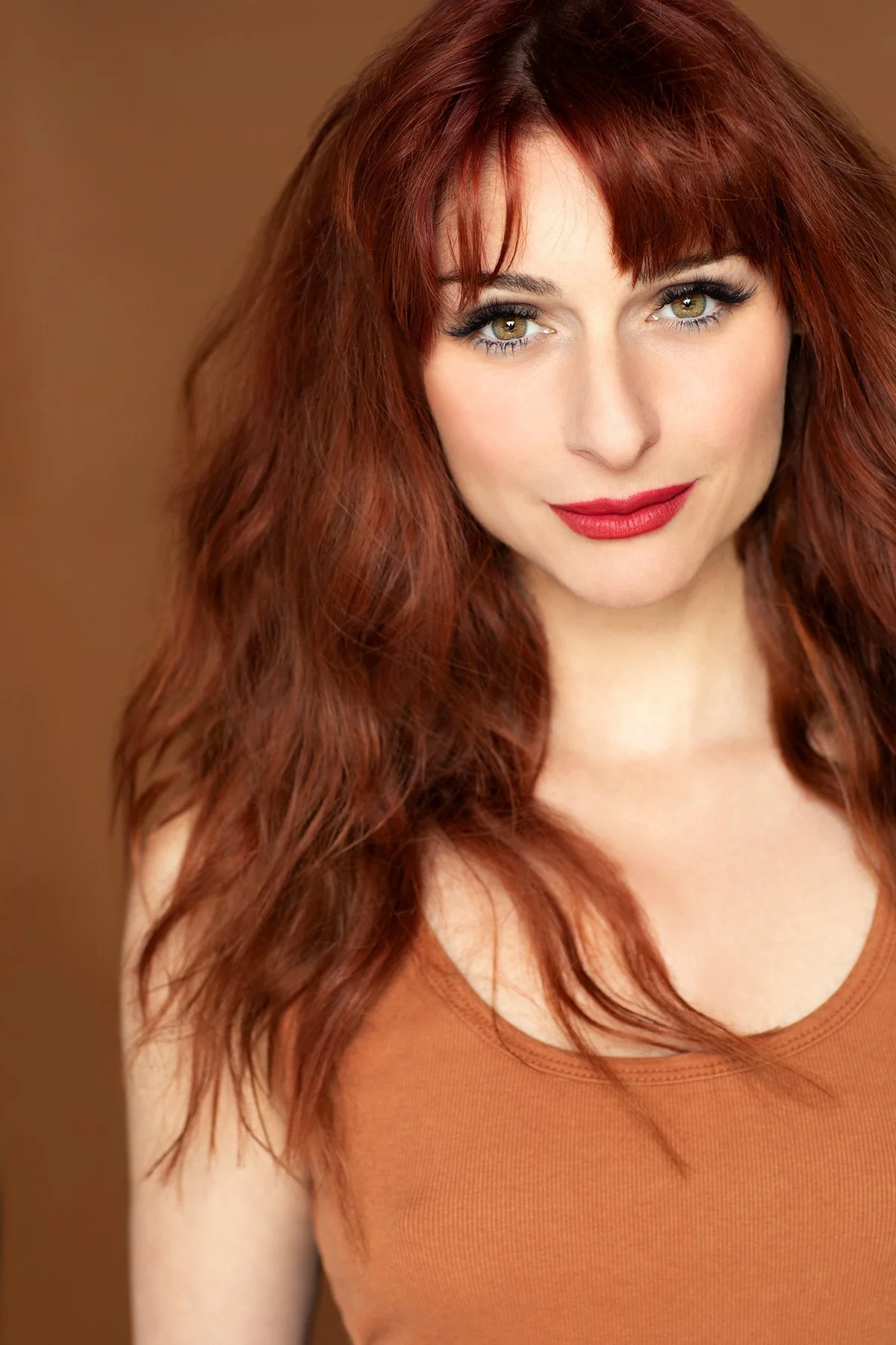 A woman with long red hair, makeup, and wearing a brown tank top, looking directly at the camera with a slight smile.