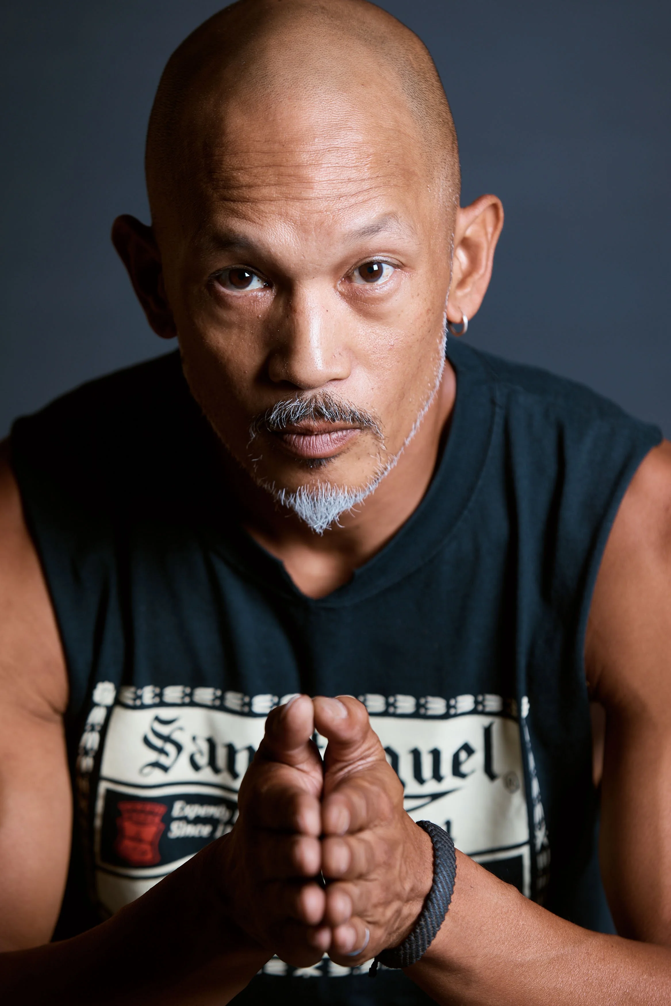 A middle-aged man with a shaved head, gray goatee, earring, wearing a black sleeveless shirt with a 'San Miguel' logo, looks directly at the camera with his hands pressed together in a prayer-like gesture against a dark background.