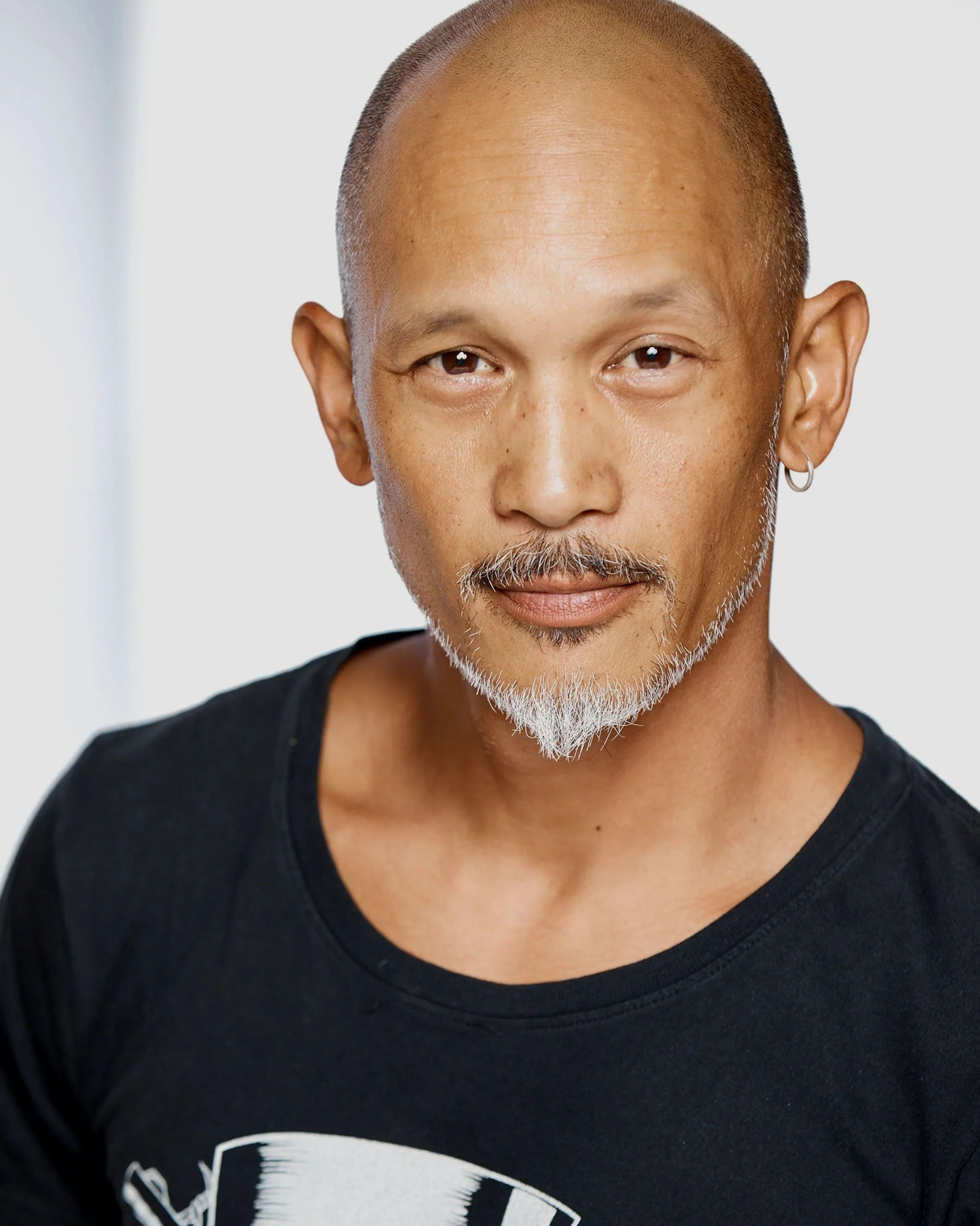 Close-up of a bald man with a gray goatee, wearing a black T-shirt and a small hoop earring in his left ear, standing against a plain light background.