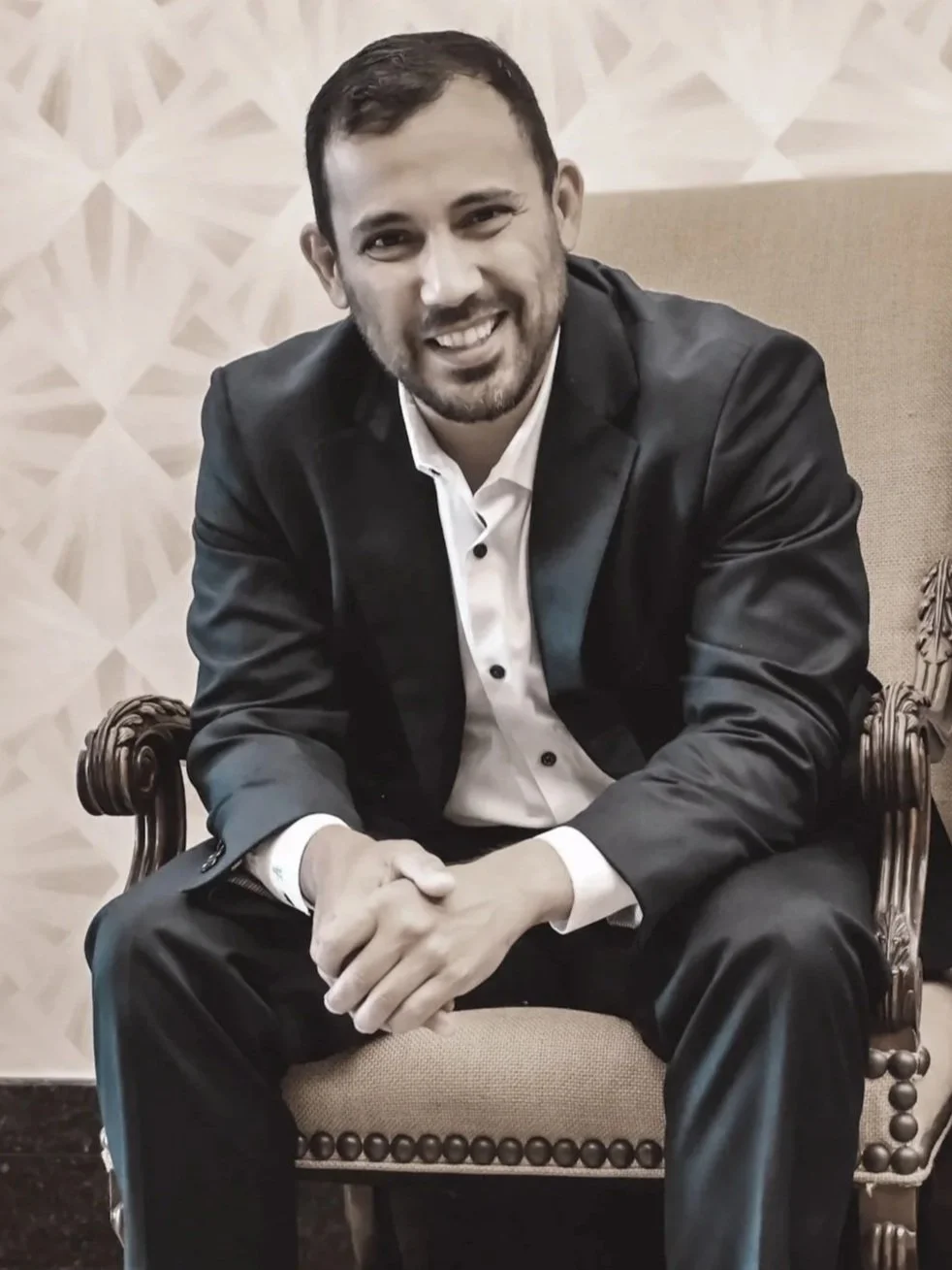 A man with dark hair and a beard wearing a black suit and white shirt, sitting on an ornate chair, smiling at the camera.