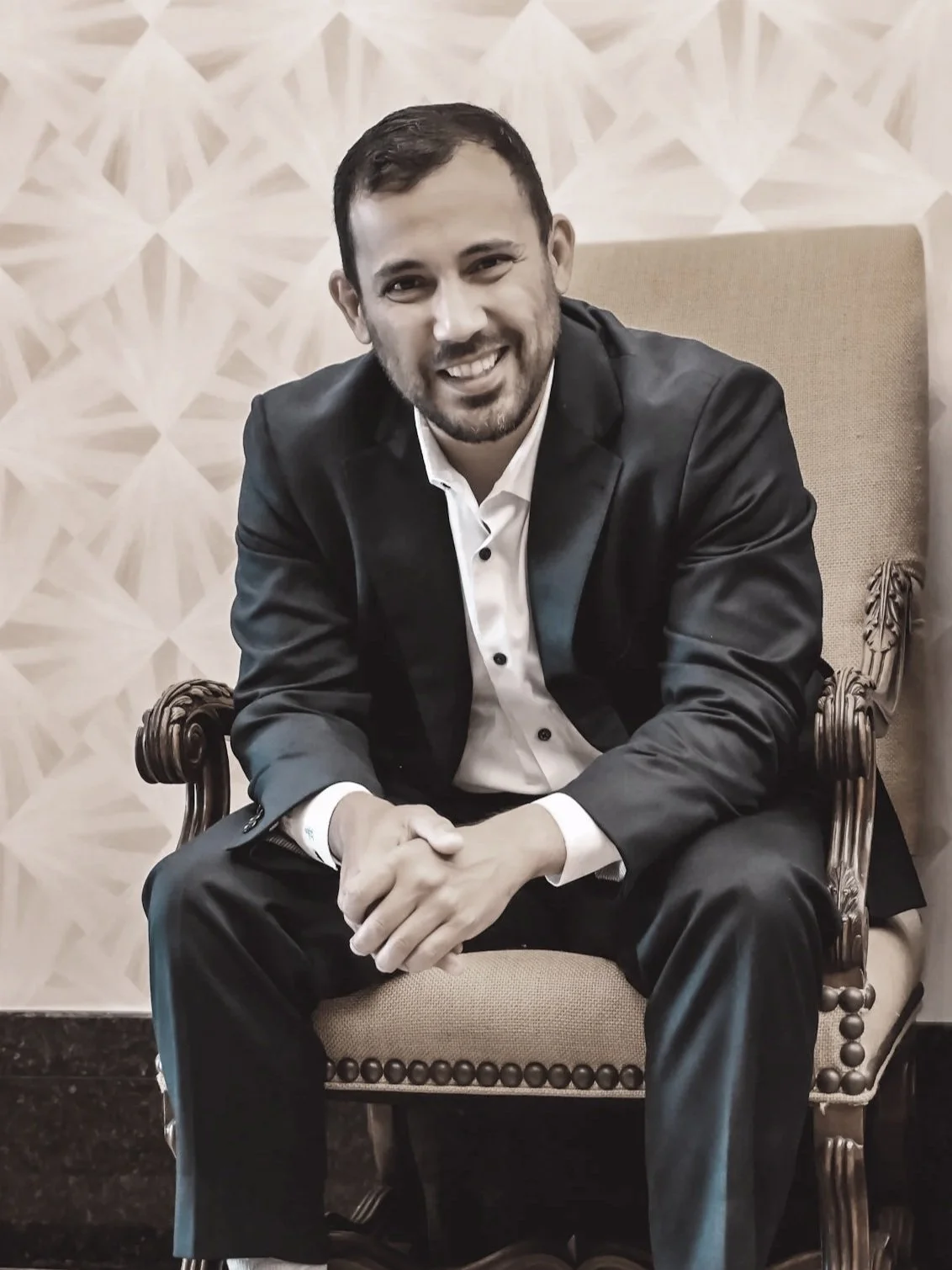 An investment advisor with dark hair and a beard, smiling, wearing a black suit jacket and white shirt, sitting on a vintage wooden armchair with beige upholstery, in front of a patterned beige wall.