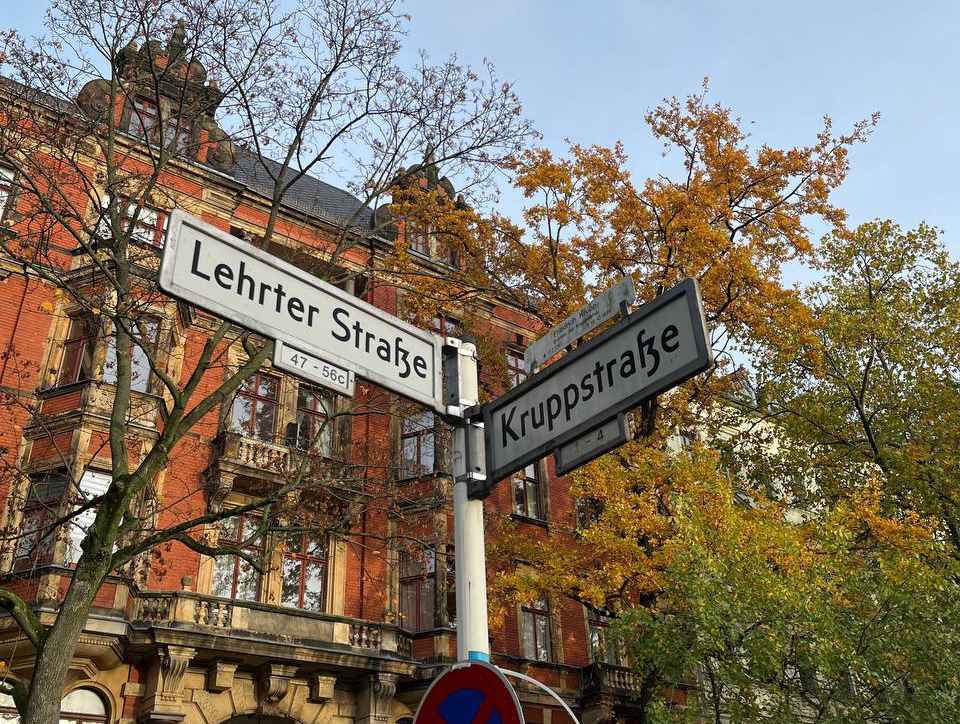 Street signs at the intersection of Lehrter Straße and Kruppstraße with a historic brick building and trees with autumn foliage in the background.