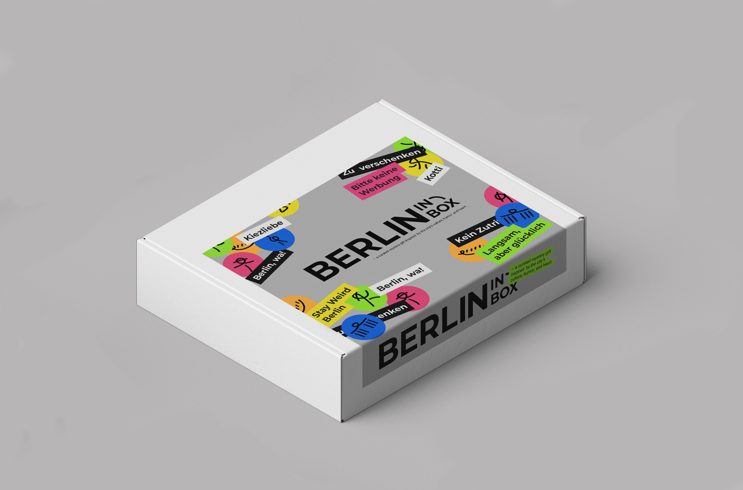 A white box with colorful stickers and the text "Berlin in a box" on the lid, featuring various tags with phrases in German, such as "Kiezliebe" and "Stay Weird Berlin, War!"